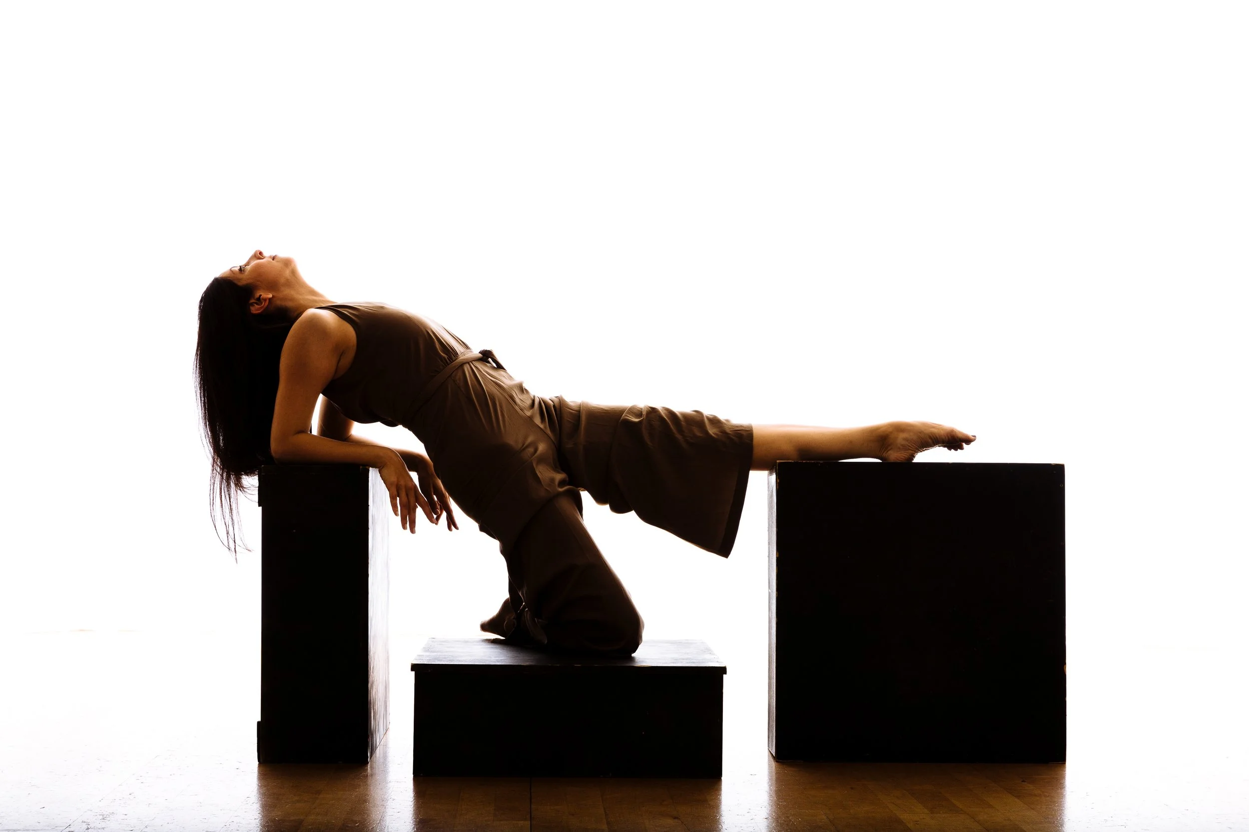 A woman in a brown dress reclining on black rectangular blocks against a white background, with her head tilted backwards and one arm resting on the block, her other hand hanging down.