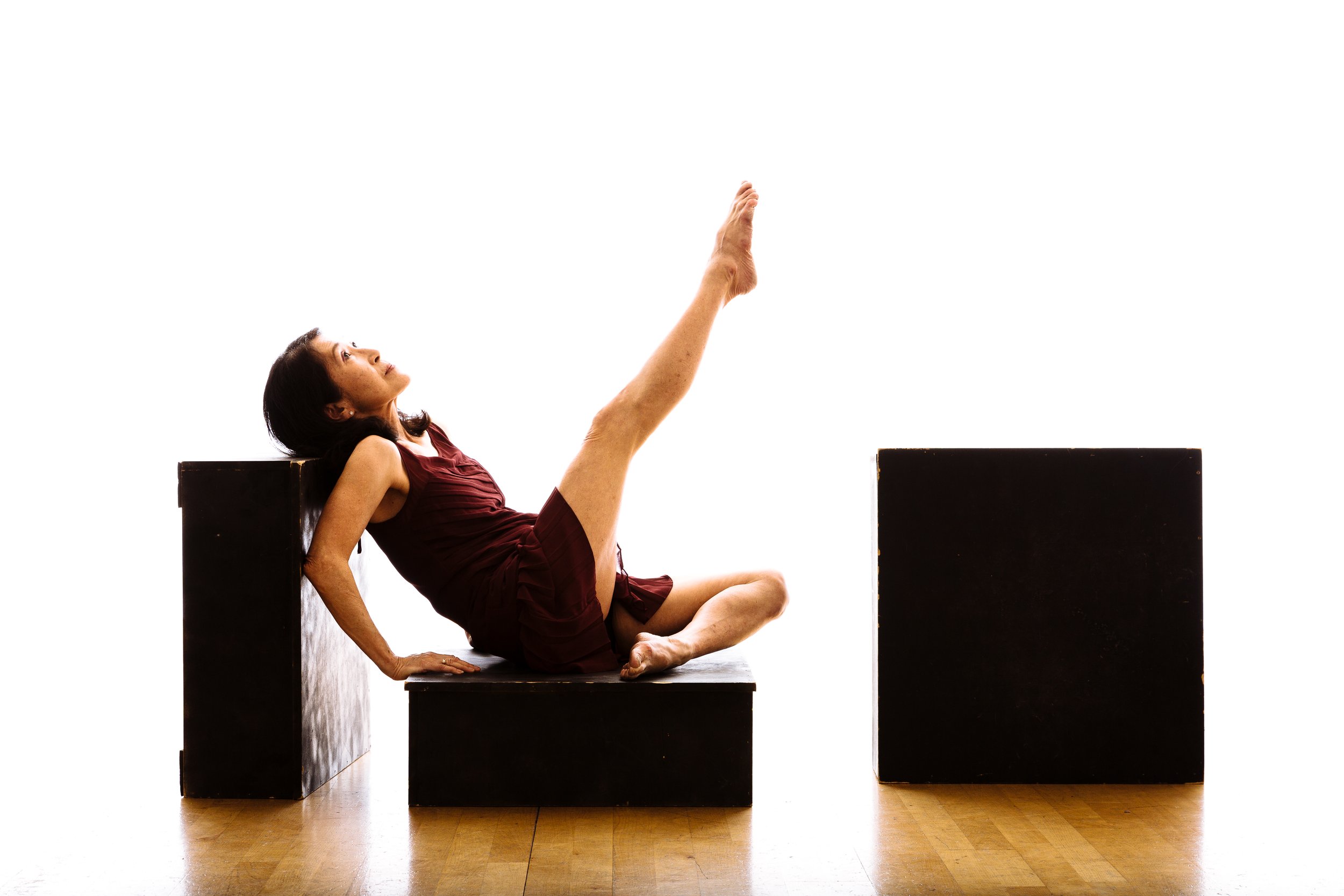 A woman in a maroon dress performing a stretching exercise or yoga pose, sitting on a black platform with one leg extended upward and her hands supporting her on the platform, against a plain white background.