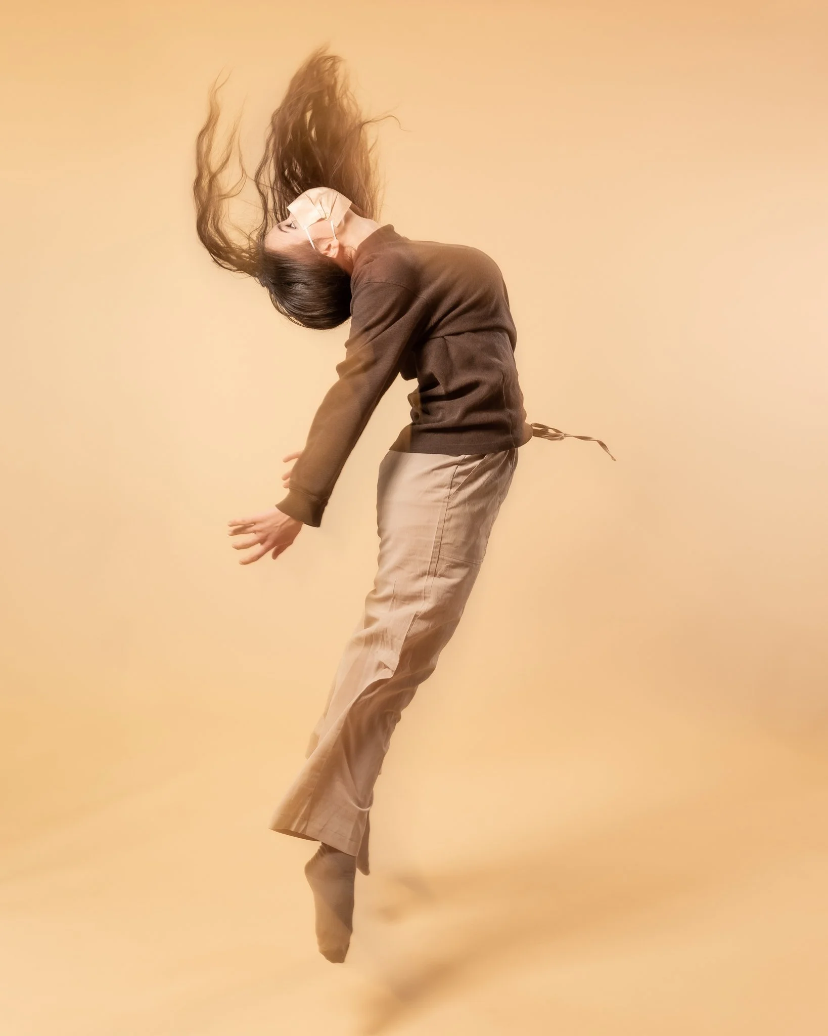A person wearing a face mask and casual clothing is captured mid-air against a beige background, with their hair flying and body bent backwards.