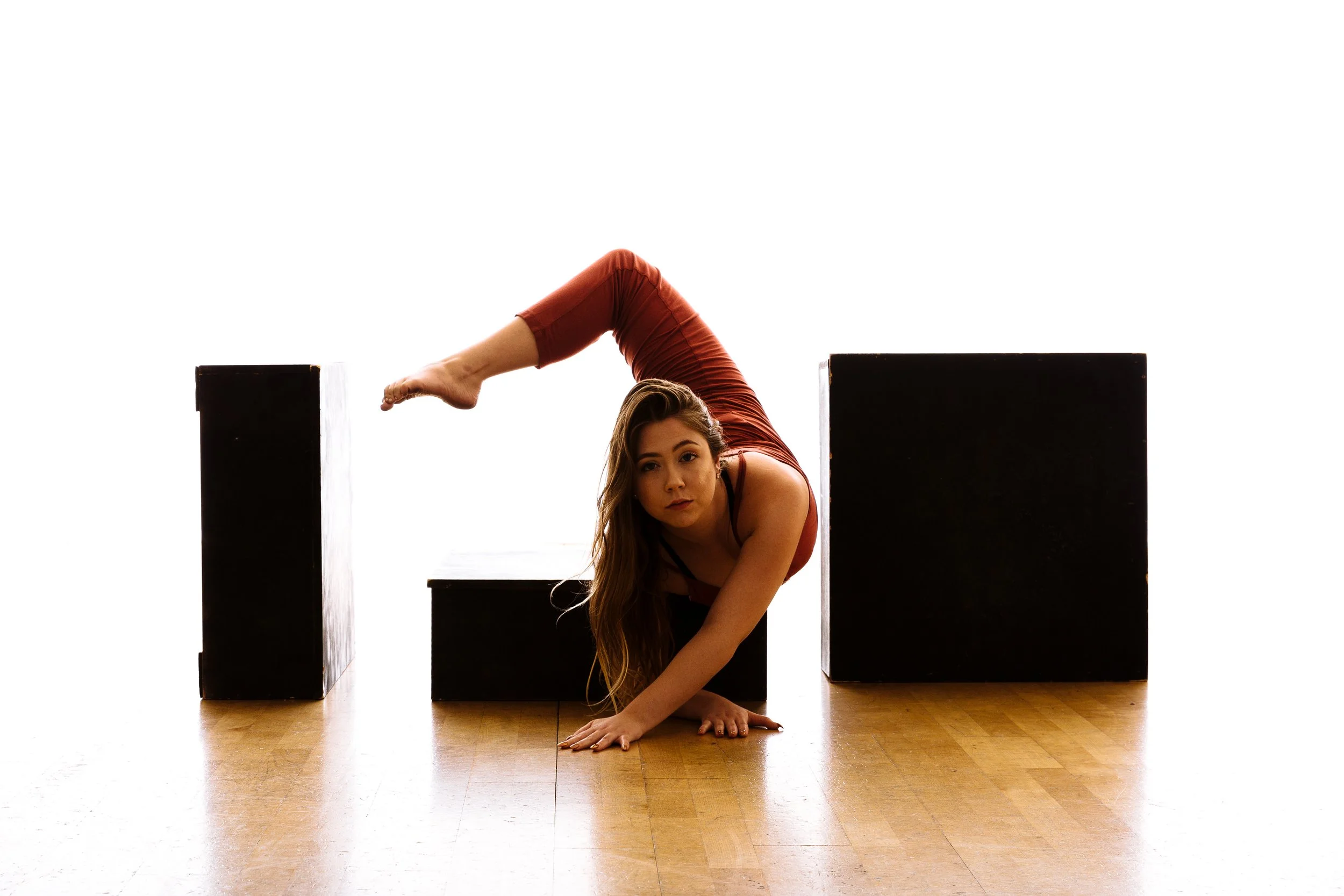 A woman in a red outfit performing a contortion pose between black rectangular blocks on a wooden floor.