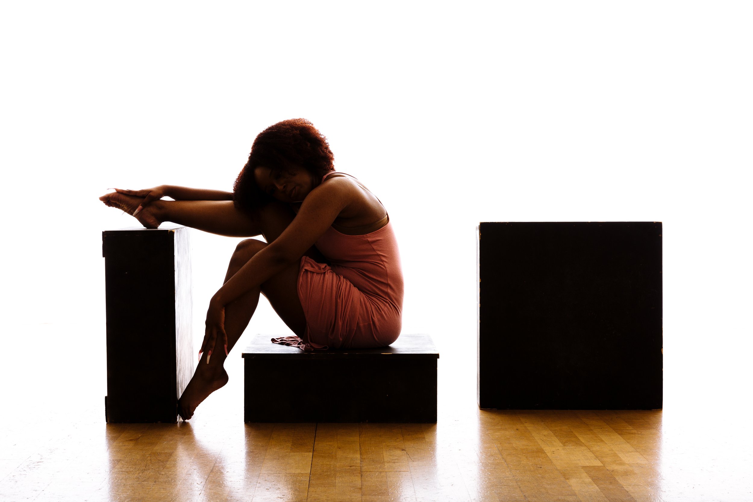 A woman in a coral dress sitting on a black box, stretching her leg over a taller black block, with her arms extended forward and her head resting on her offset knee, against a bright white background.