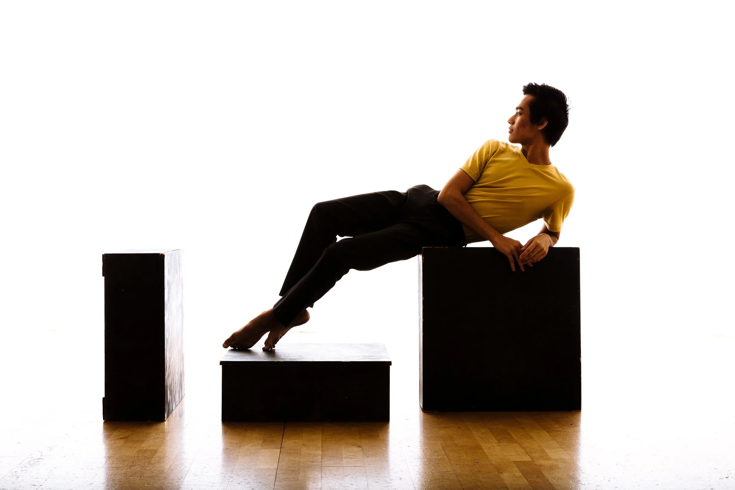 A person with short dark hair, wearing a yellow shirt and black pants, is reclining on black rectangular blocks against a white background.