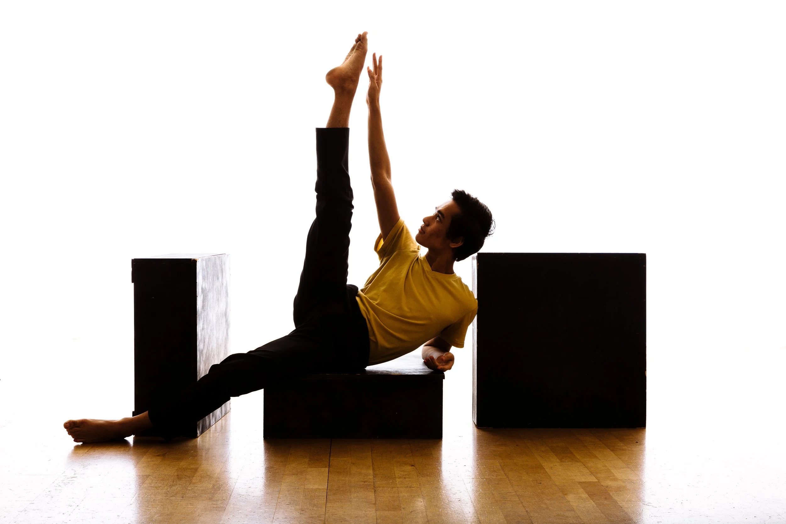 A person in a yellow shirt and black pants performing a stretching exercise on black blocks against a white background.