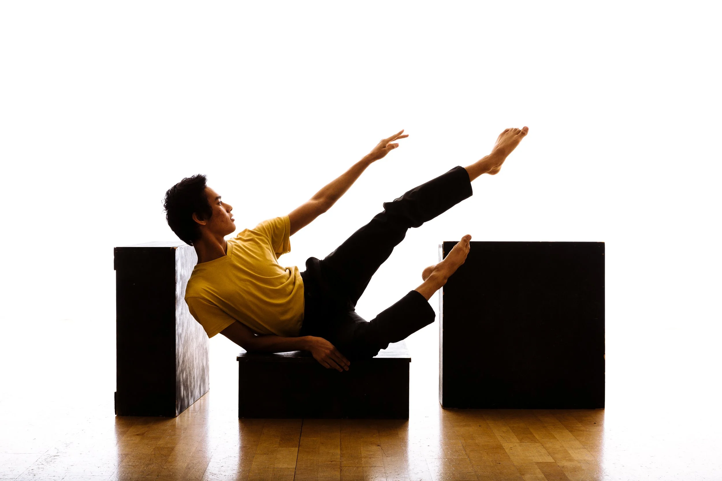 Person in a yellow shirt and black pants is doing a yoga pose or exercise on black blocks on a wooden floor, with a white background.