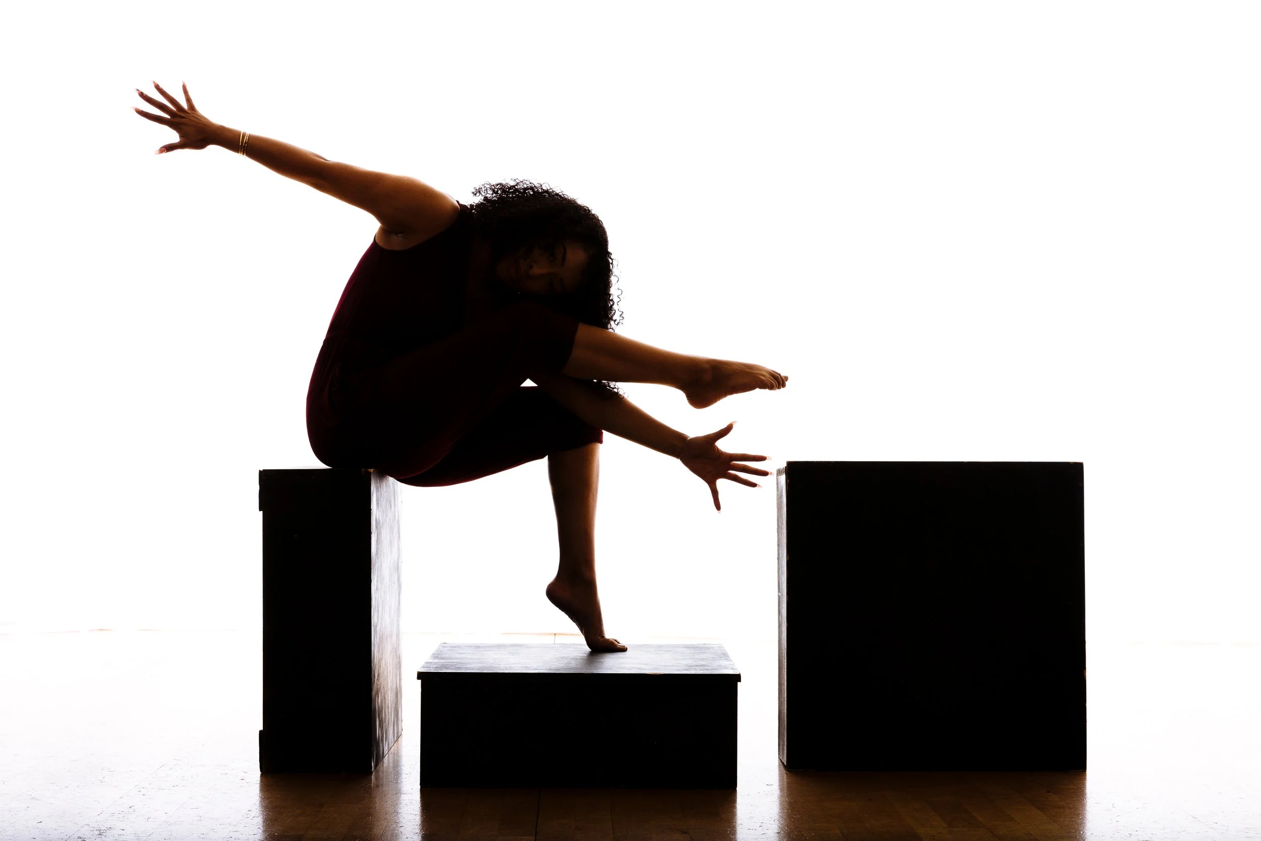 A woman with curly hair balancing on a platform with her arms outstretched, in a dance or yoga pose, silhouetted against a white background.