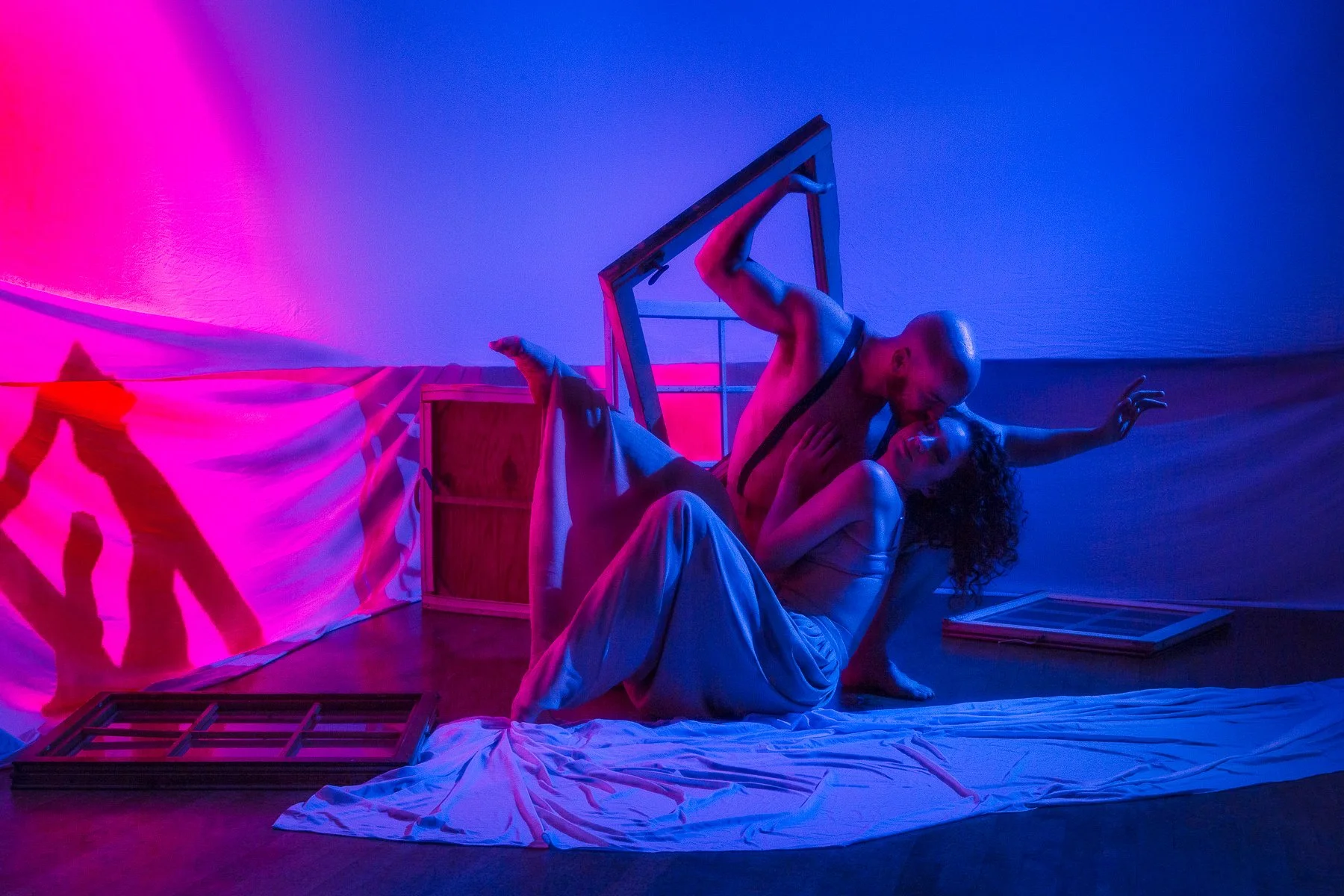 A man and woman in a dance pose on the floor with colorful pink, purple, and blue lighting, with props and fabrics around them.