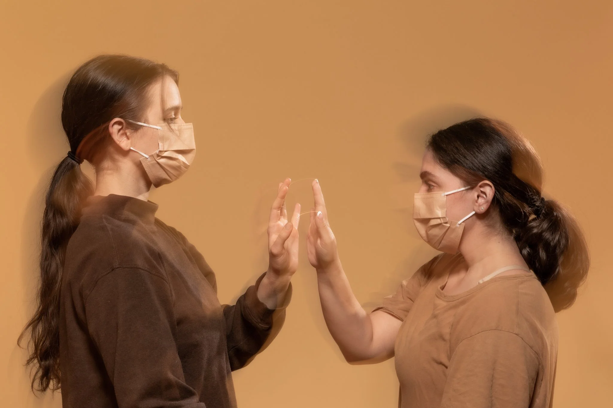 Two women wearing brown face masks and casual brown clothing face each other, with their hands raised and fingers touching, against a plain tan background.