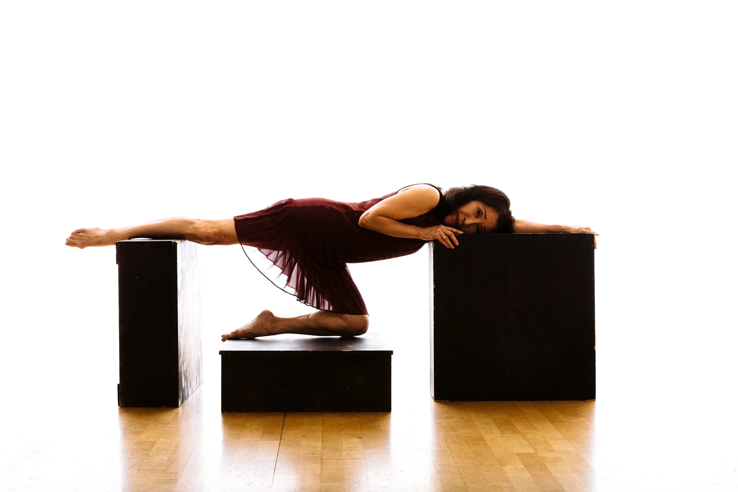 A woman in a maroon dress performing a contemporary dance pose on black cube platforms against a plain white background.