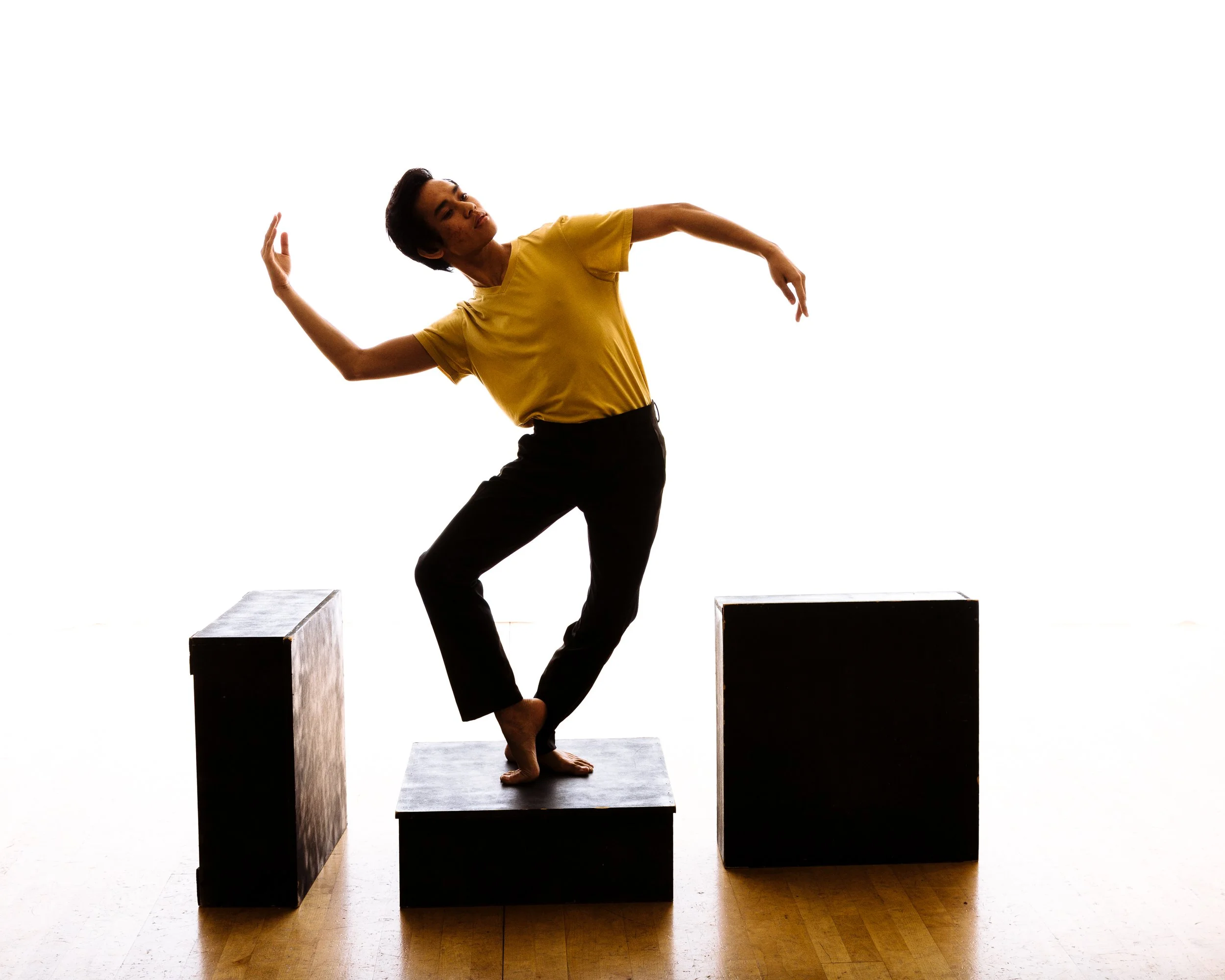 Person in yellow shirt and black pants balancing on a black block, with two other black blocks nearby, against a white background.