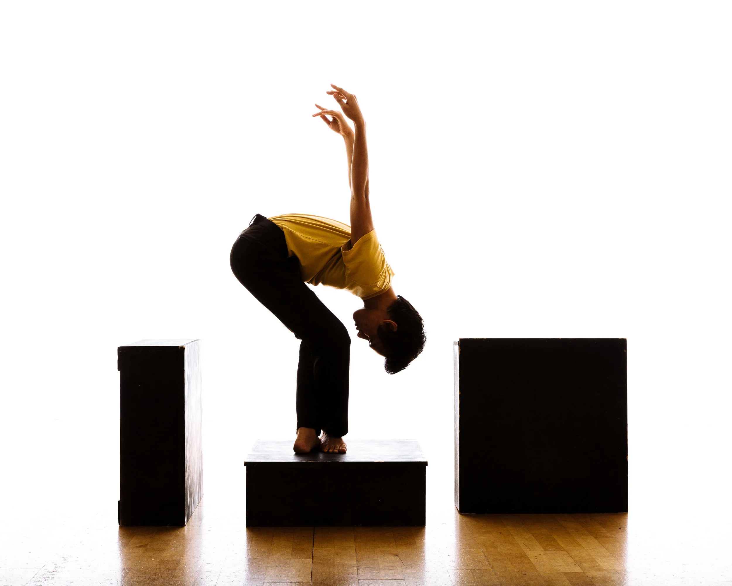 Person in a yellow shirt and black pants performing a forward bend yoga pose on a black platform with two black blocks on either side.