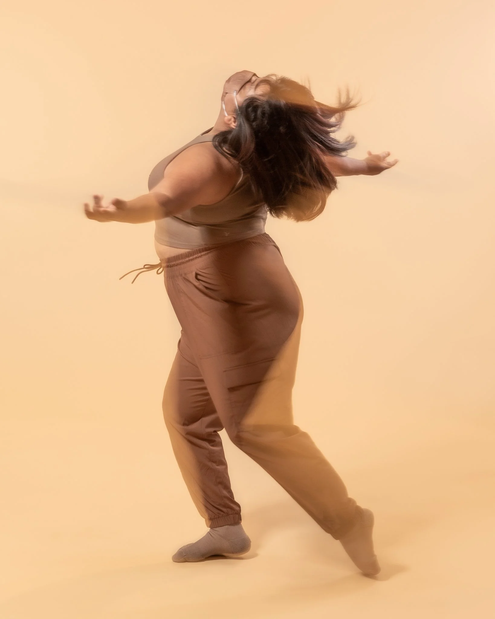 A person dancing with long hair flowing, wearing a brown tank top and brown sweatpants against a beige background.