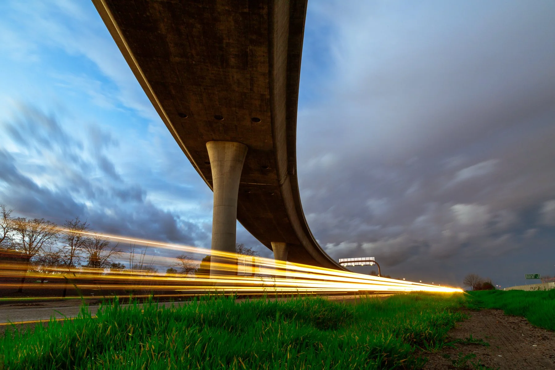 Low-angle view of an overpass bridge at dusk with light trails from passing cars on the road below.