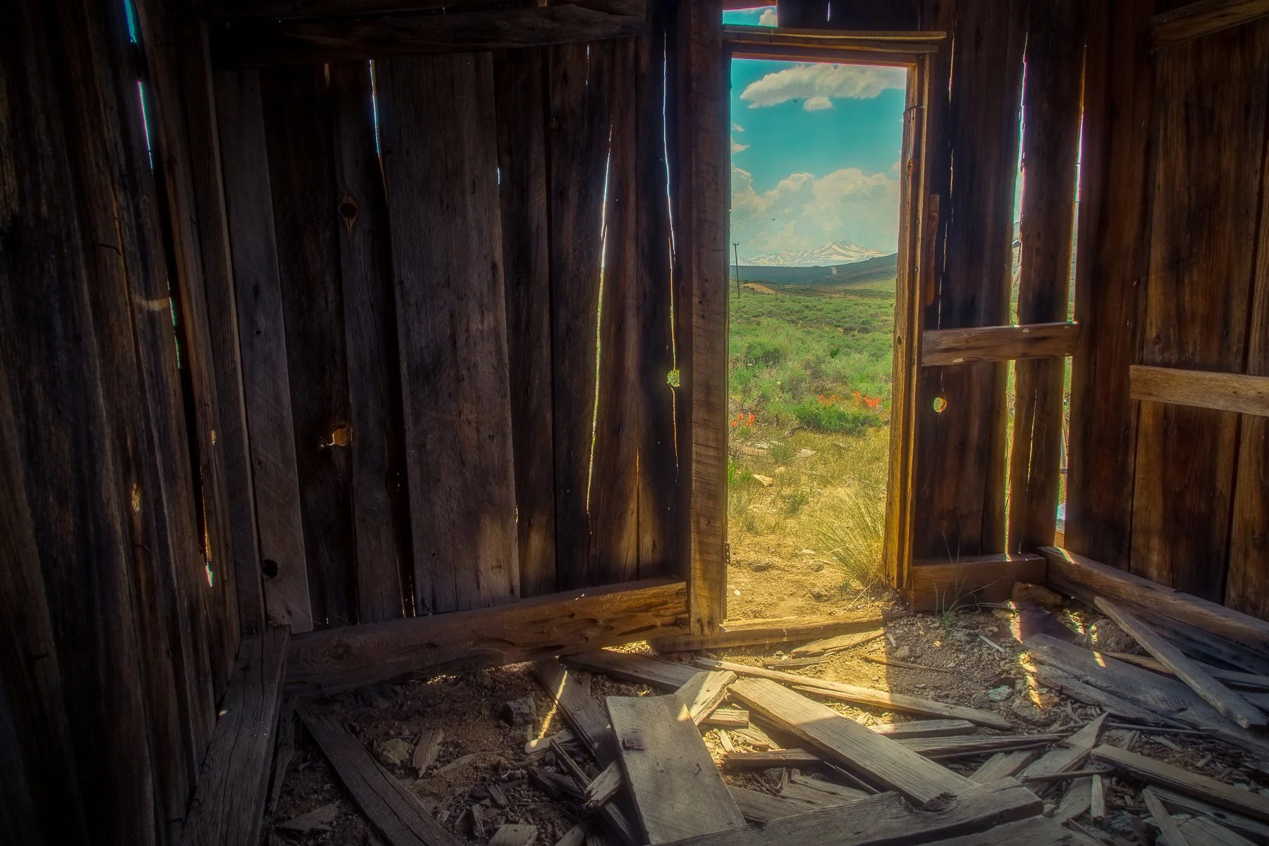 Interior of an abandoned wooden building with broken floorboards, overlooking a green landscape with mountains in the distance, under a partly cloudy sky.
