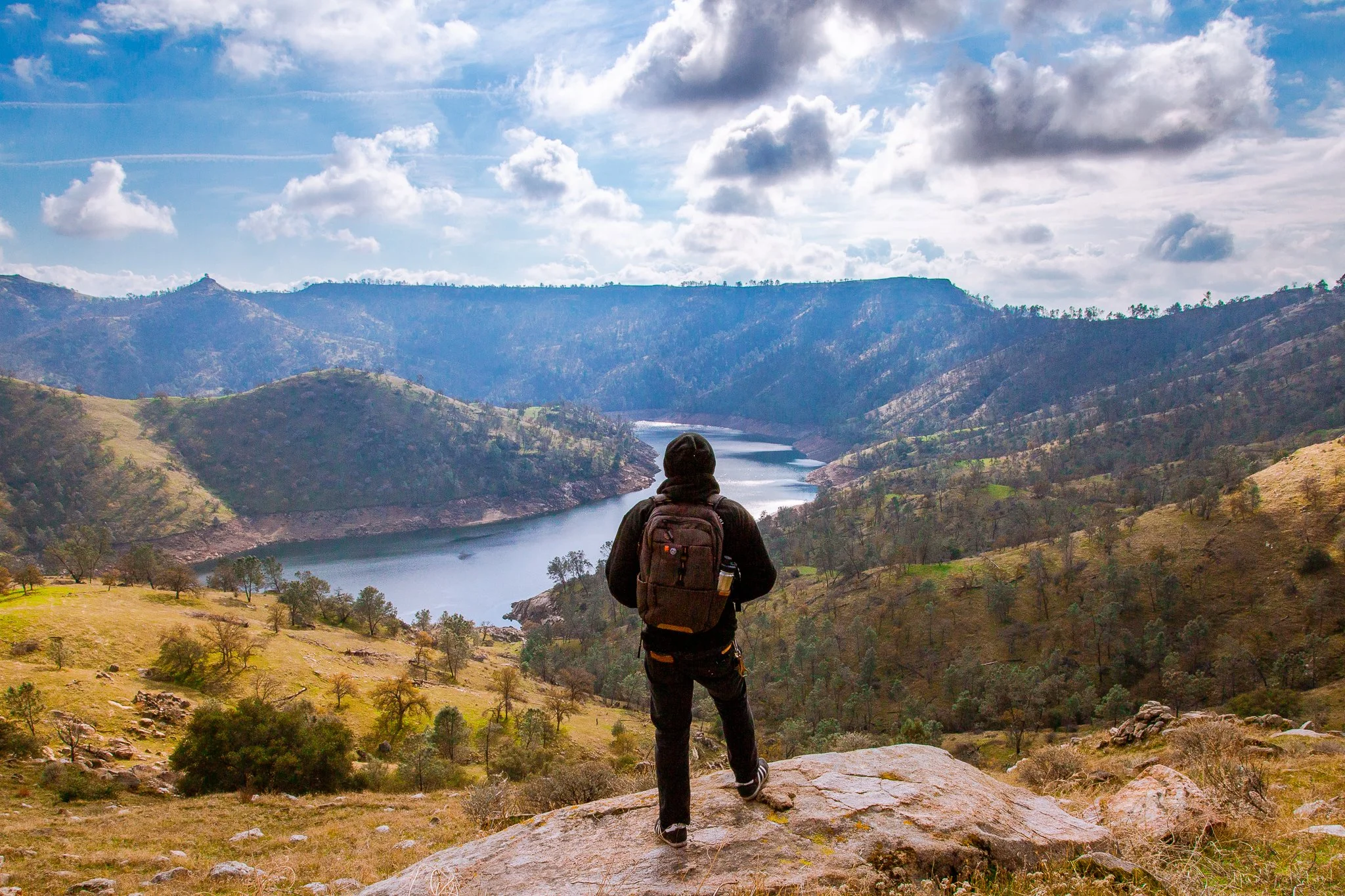 A person with a backpack stands on a rock overlooking a mountainous landscape and a river valley under partly cloudy skies.