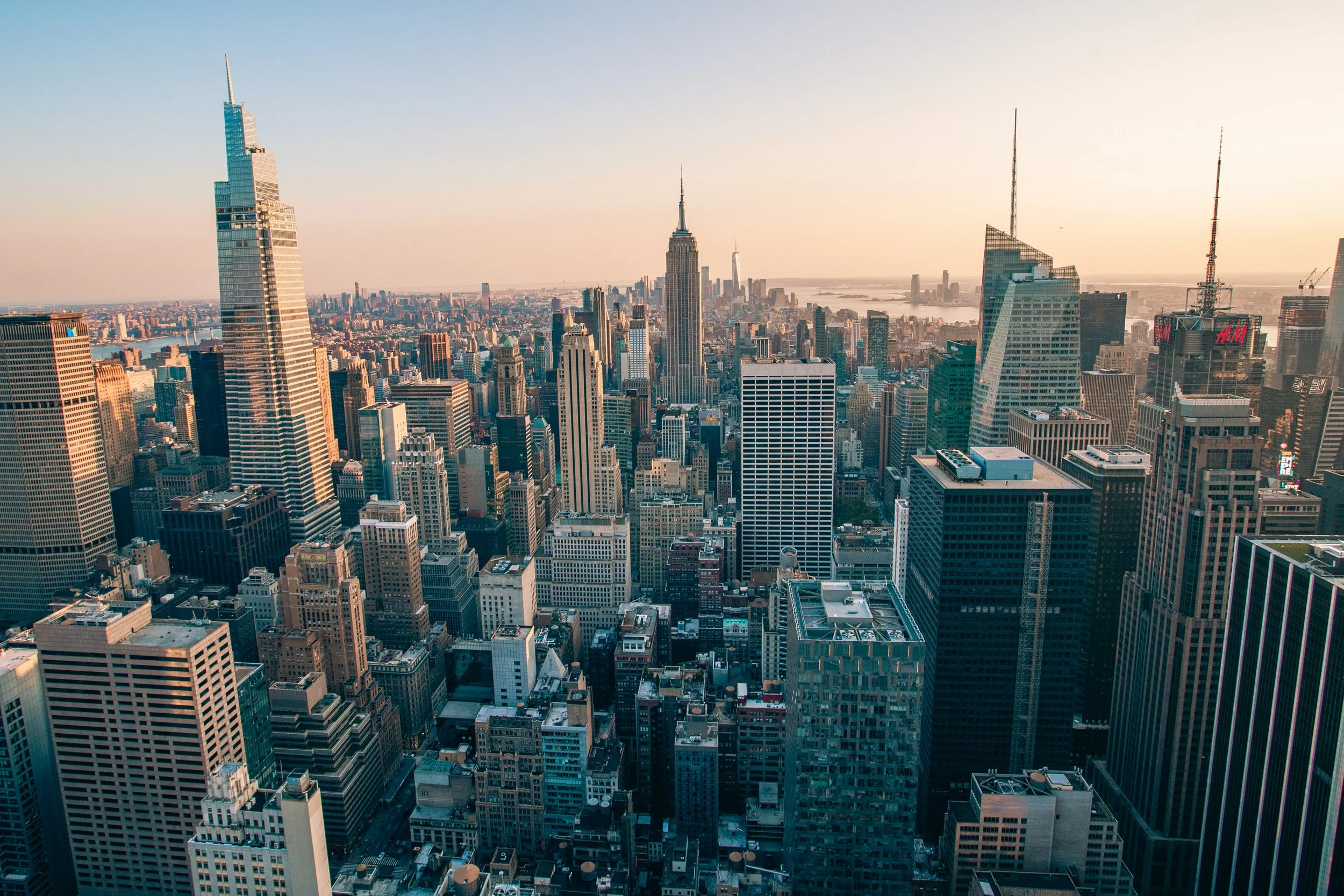 Skyline of New York City at sunset with numerous tall skyscrapers, including the Empire State Building in the center, and the city skyline extending to the horizon.