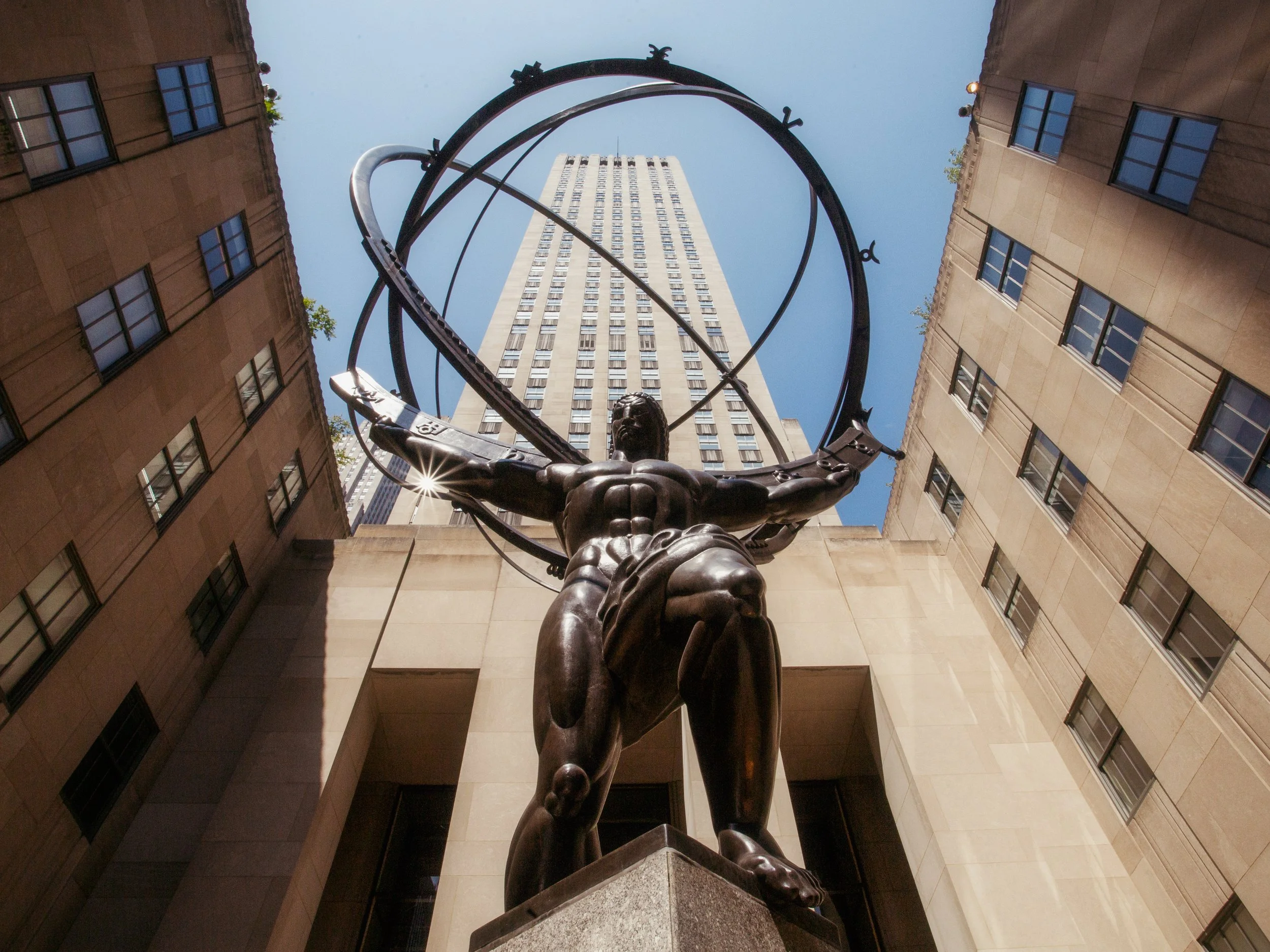 Low-angle view of a statue of Atlas holding a globe with the Empire State Building in the background, around a courtyard in New York City.