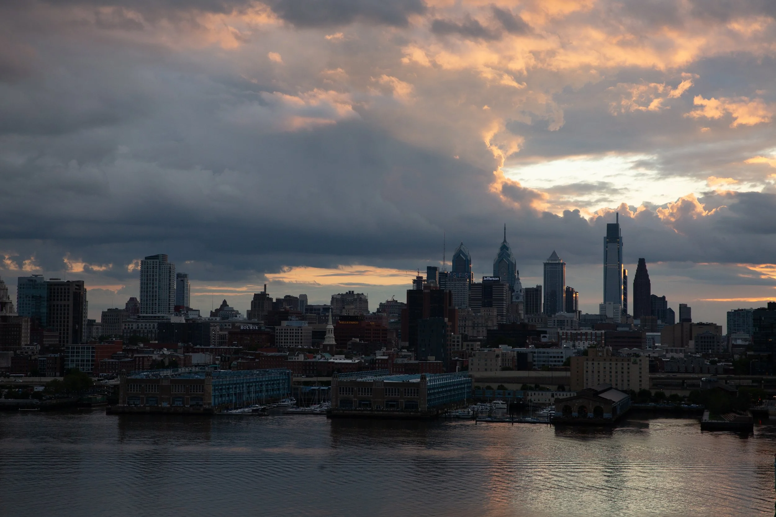 Philadelphia skyline at sunset with clouds overhead and water in the foreground.