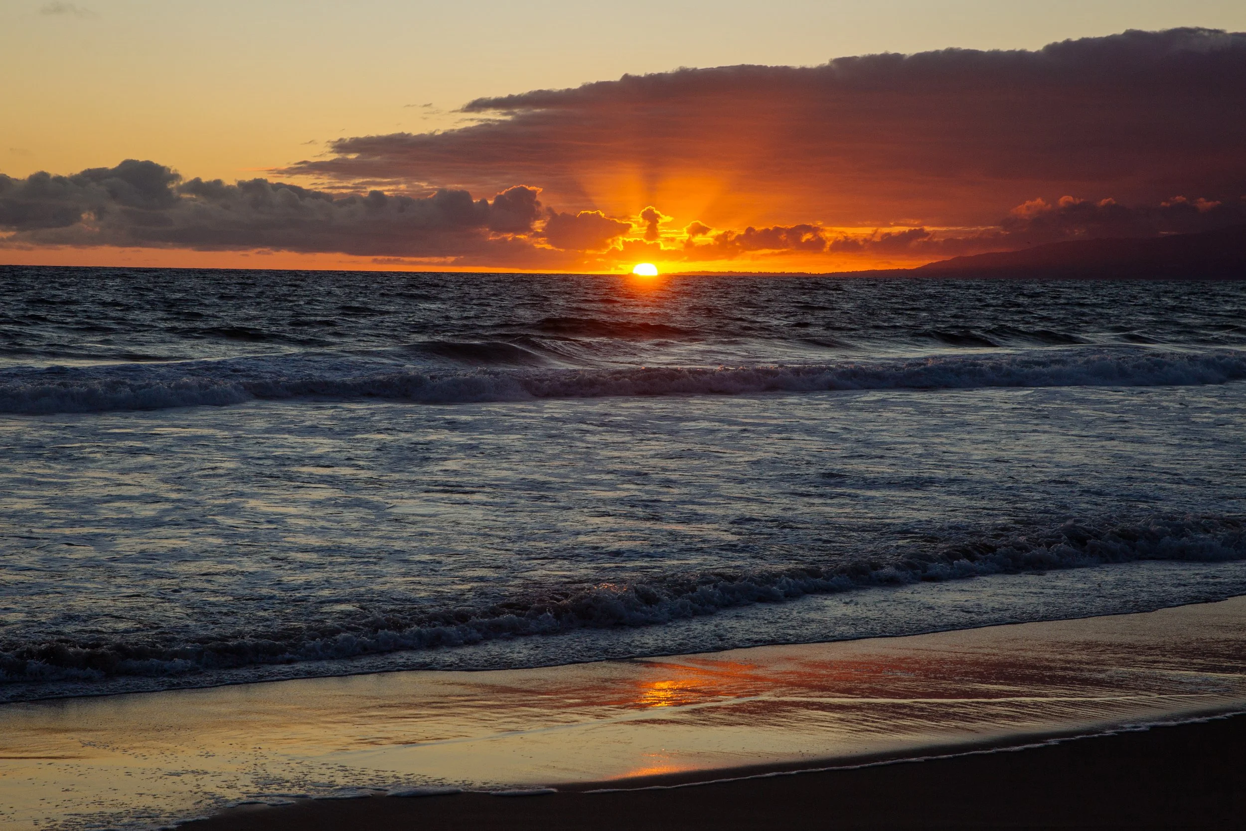A sunset over the ocean with the sun near the horizon, casting orange and yellow hues in the sky and reflecting on the water, with clouds overhead.