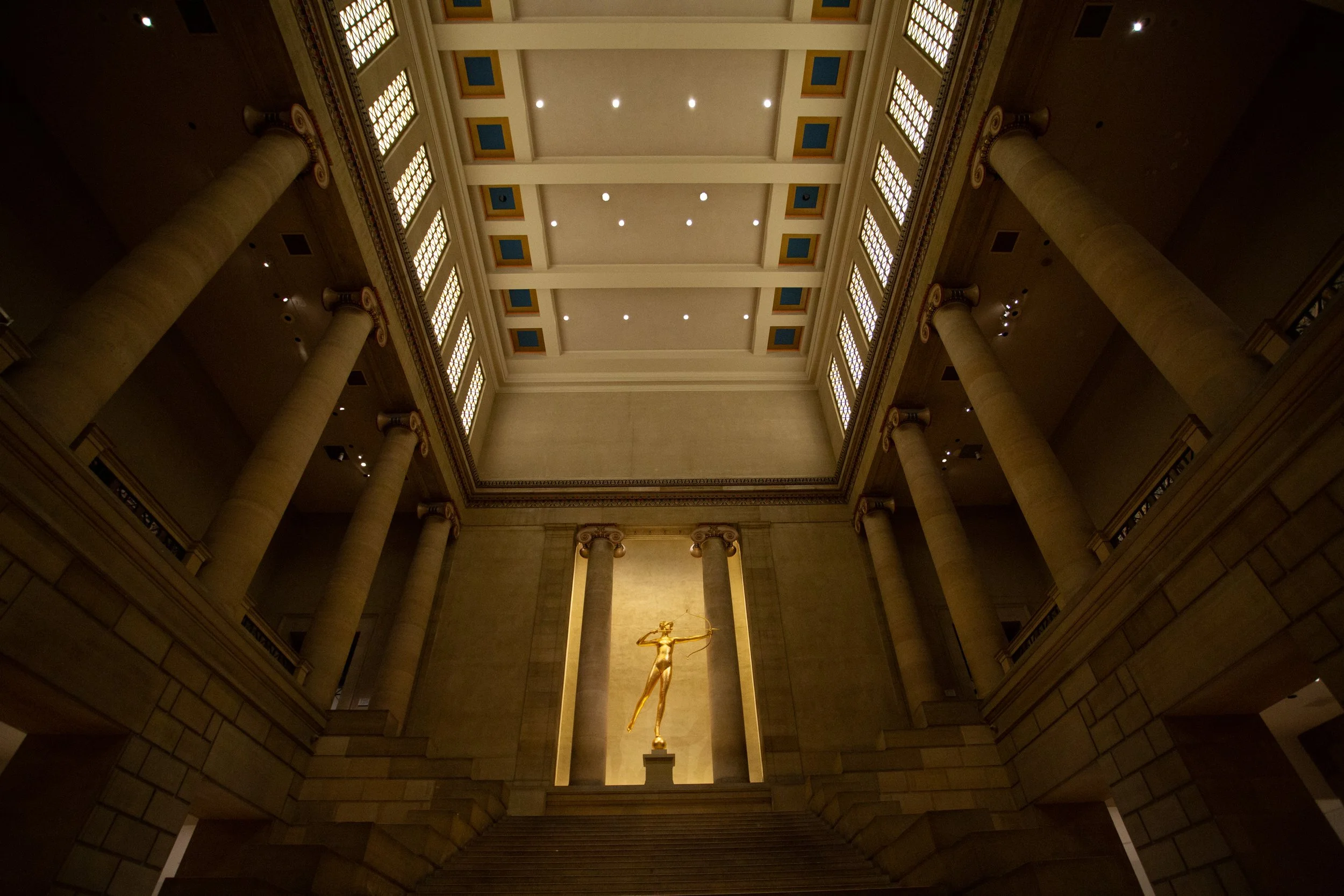 Interior view of a grand building with tall columns, a staircase leading up to a golden statue of a woman holding a bow, and a high ceiling with ornate molding and lights.