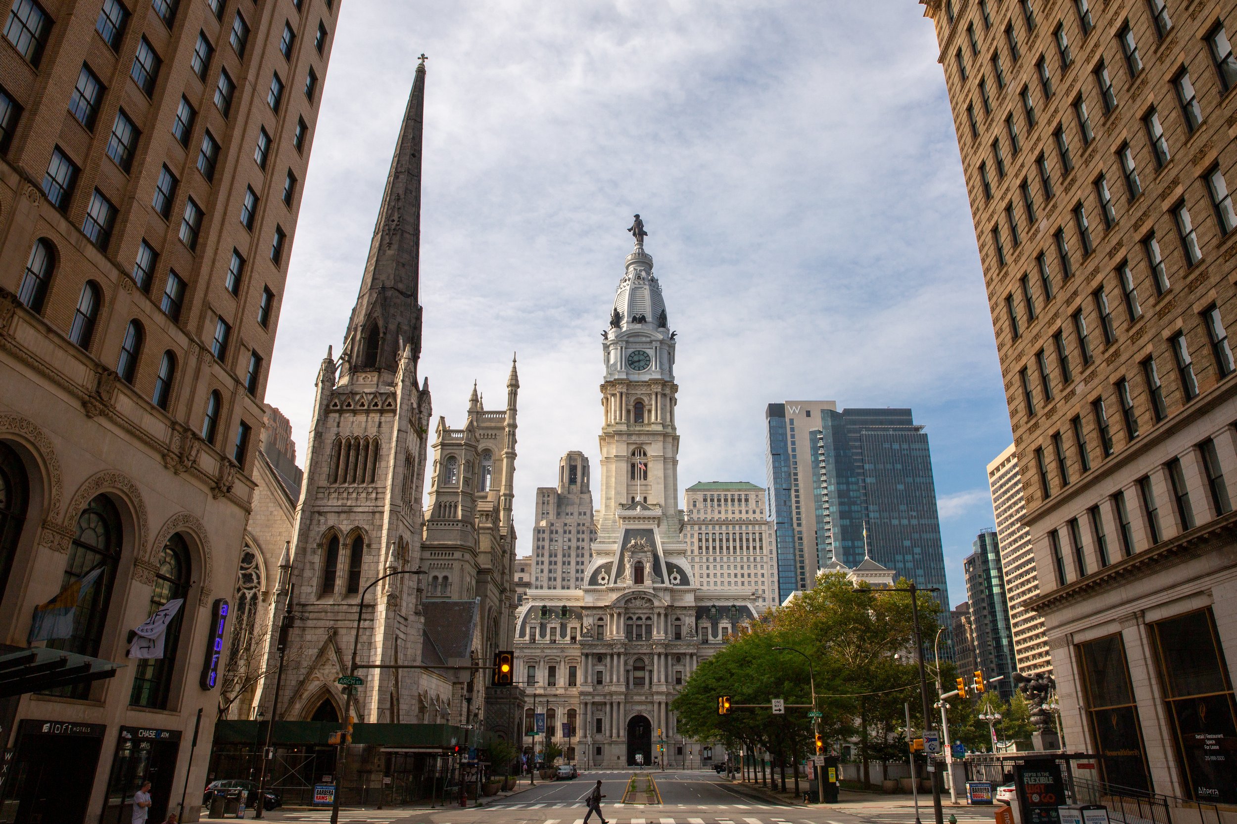 Cityscape featuring a historic clock tower and church, surrounded by modern high-rise buildings, with trees and a street in the foreground.