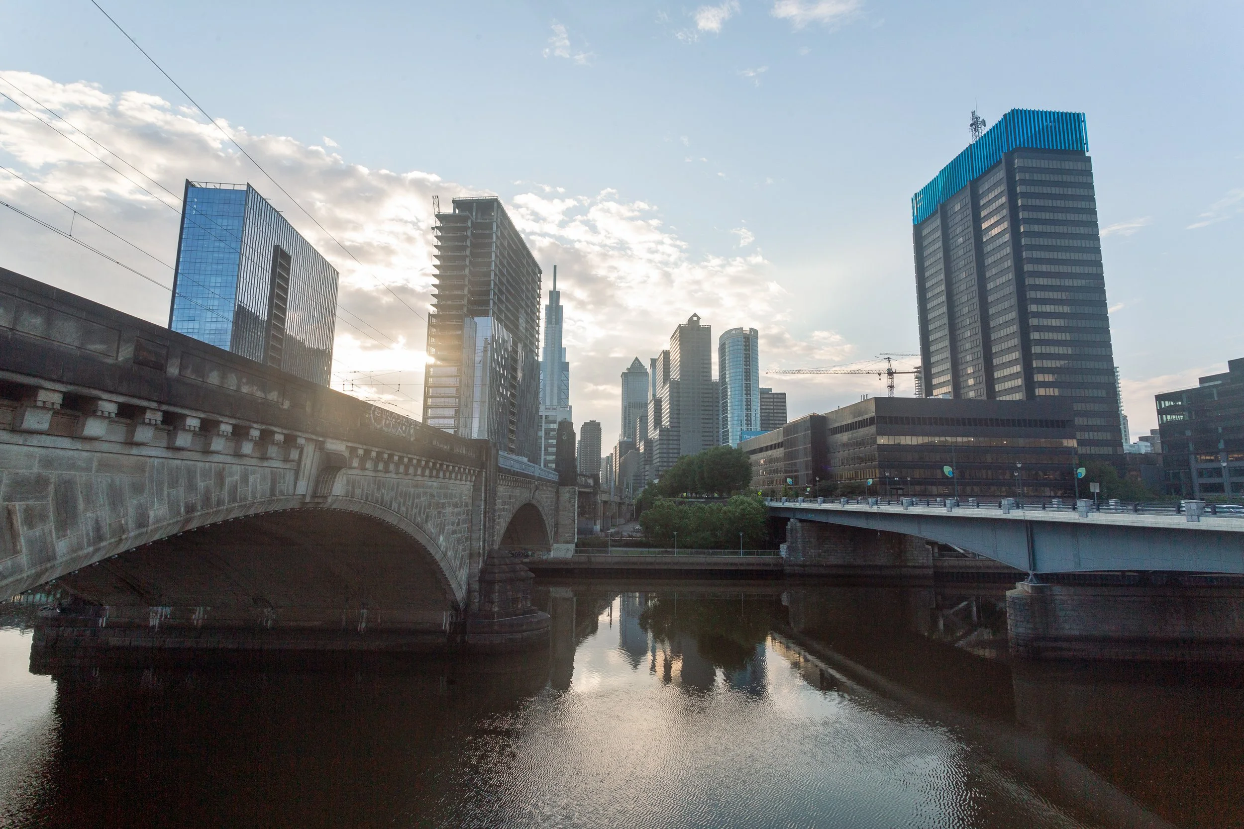City skyline with modern skyscrapers, a bridge over a river, and a partly cloudy sky during sunset.