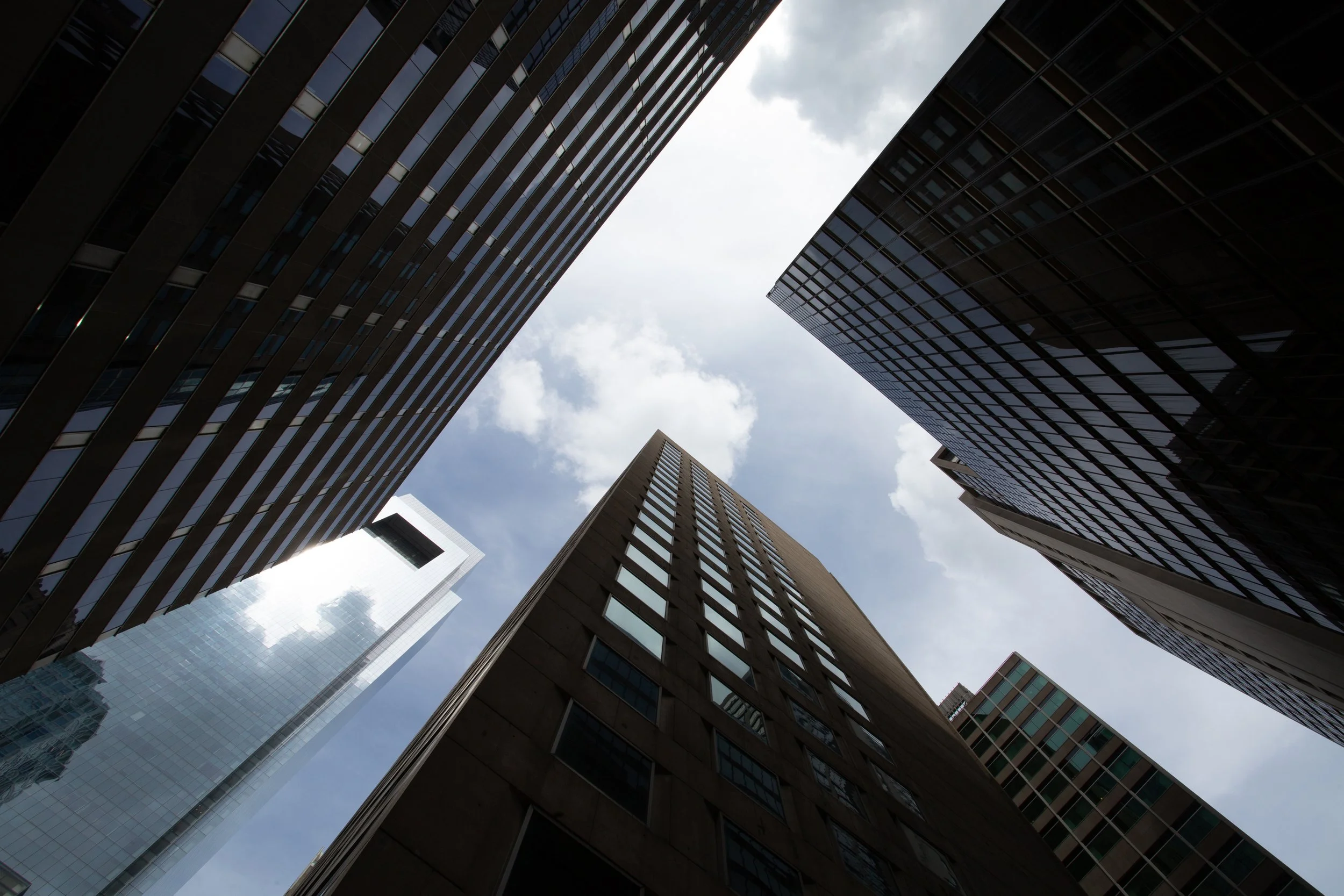 Looking up at tall modern skyscrapers with glass and concrete facades, converging towards a cloudy sky.
