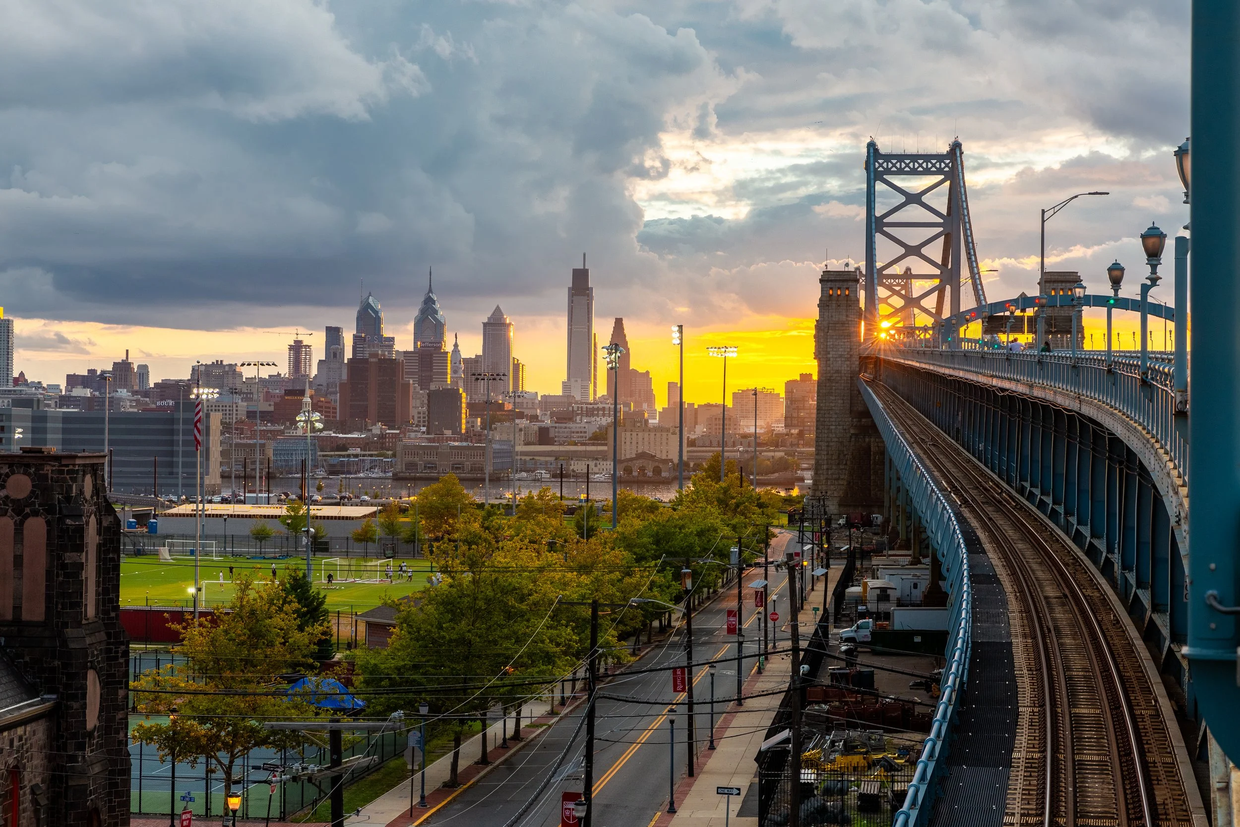 Sunset over Philadelphia skyline with Benjamin Franklin Bridge in the foreground, a park with tennis courts, and a street below.