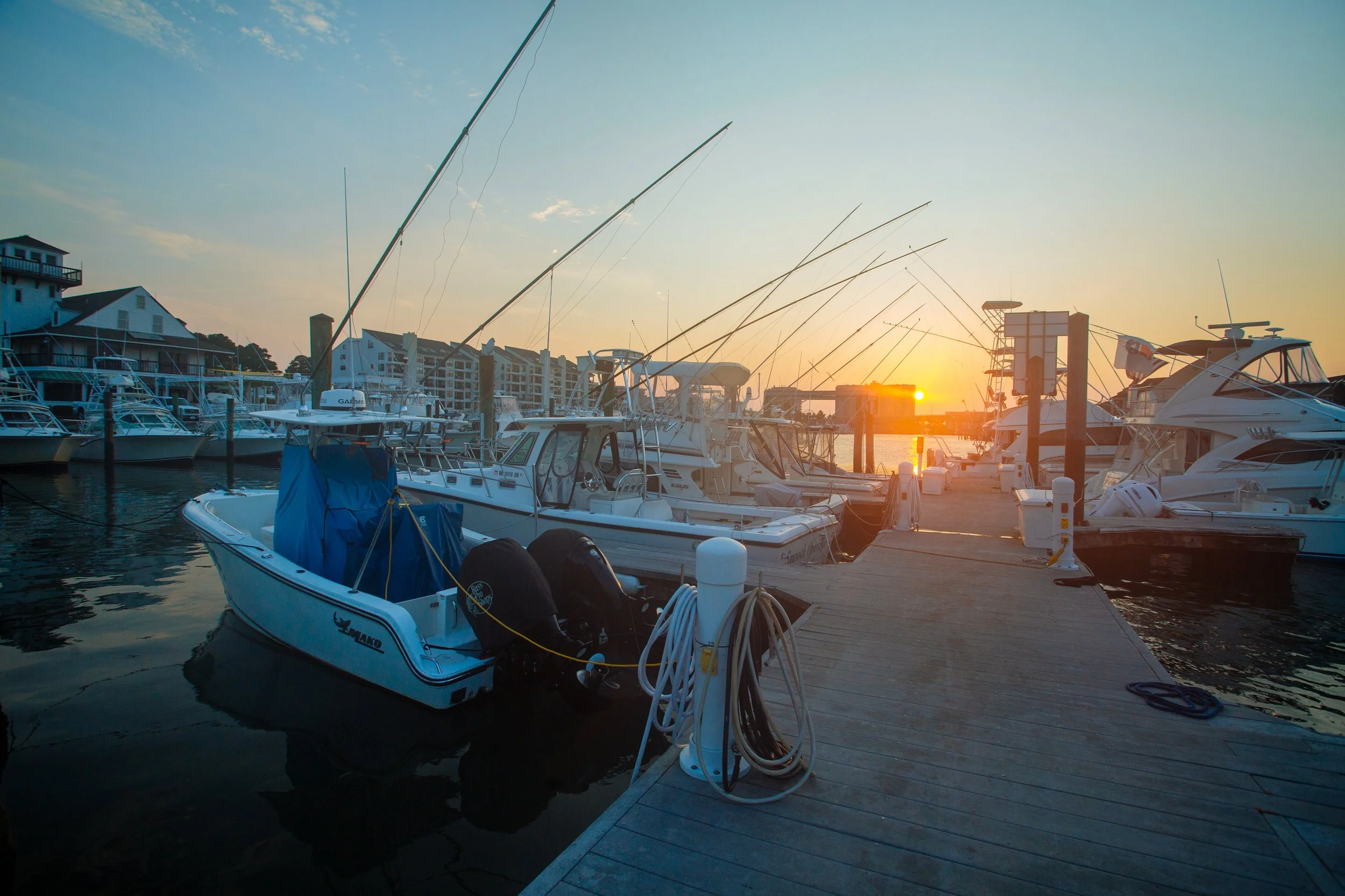 A marina at sunset with boats docked along a wooden pier, the sun low on the horizon casting a warm glow over the scene.