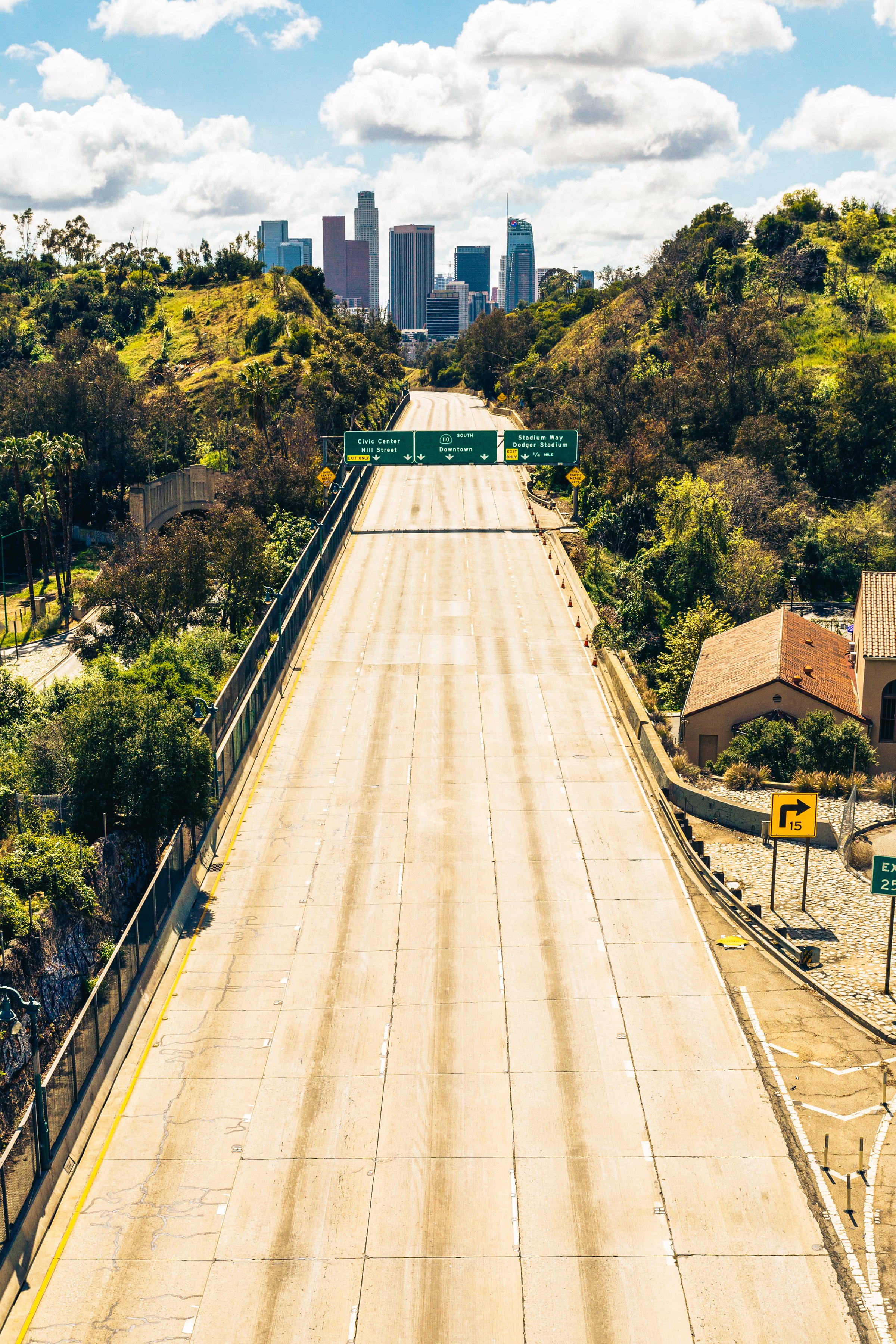 Empty freeway with green signs and downtown Los Angeles skyline in the background, overcast sky with clouds.
