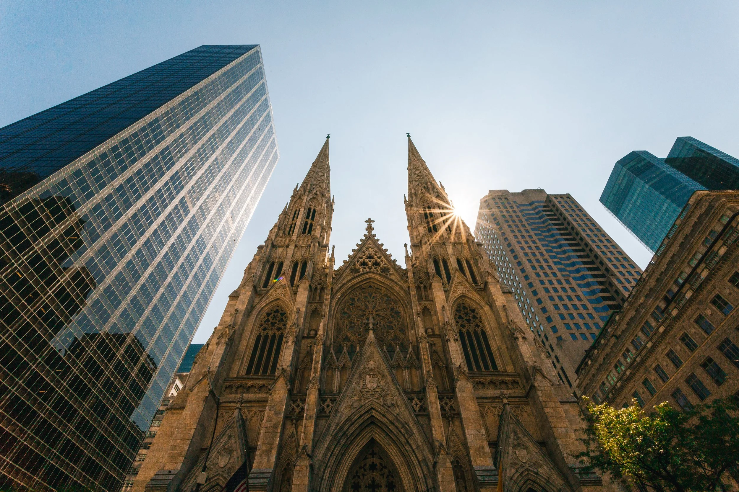 A Gothic-style church with two tall spires surrounded by modern skyscrapers, with the sun shining through the spires.