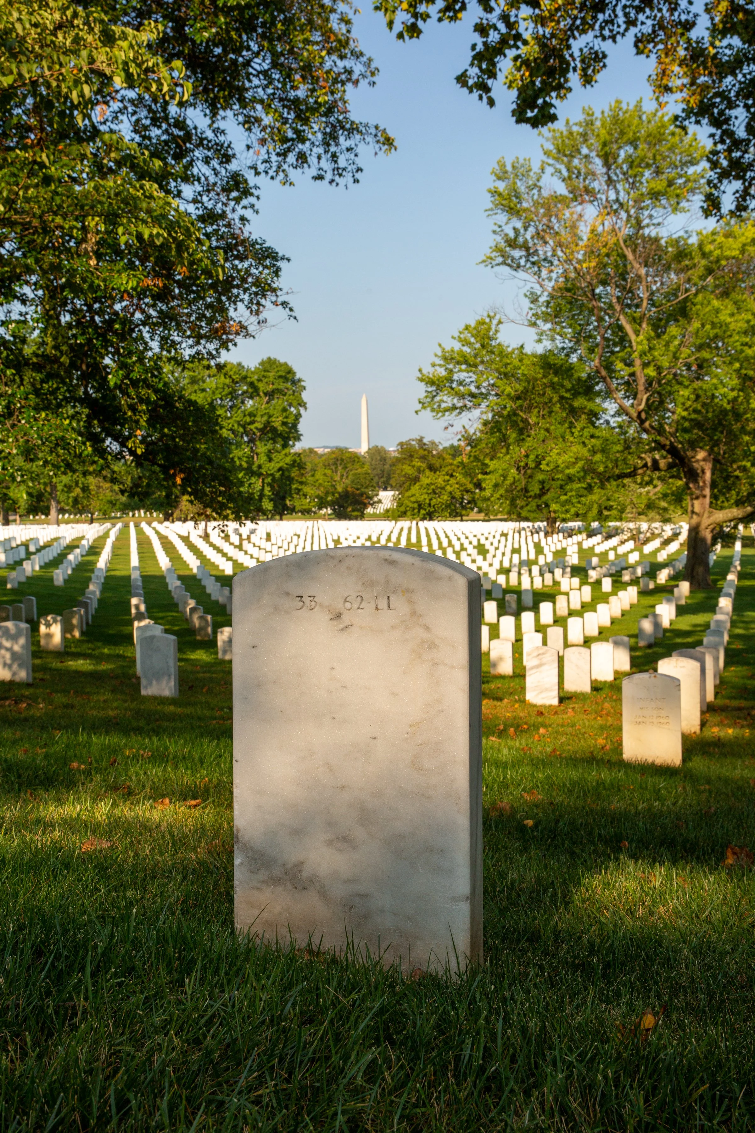 A white marble grave marker in Arlington National Cemetery with numerous rows of similar white markers stretching into the distance. Green trees line the cemetery, and the Washington Monument is visible in the background against a clear blue sky.