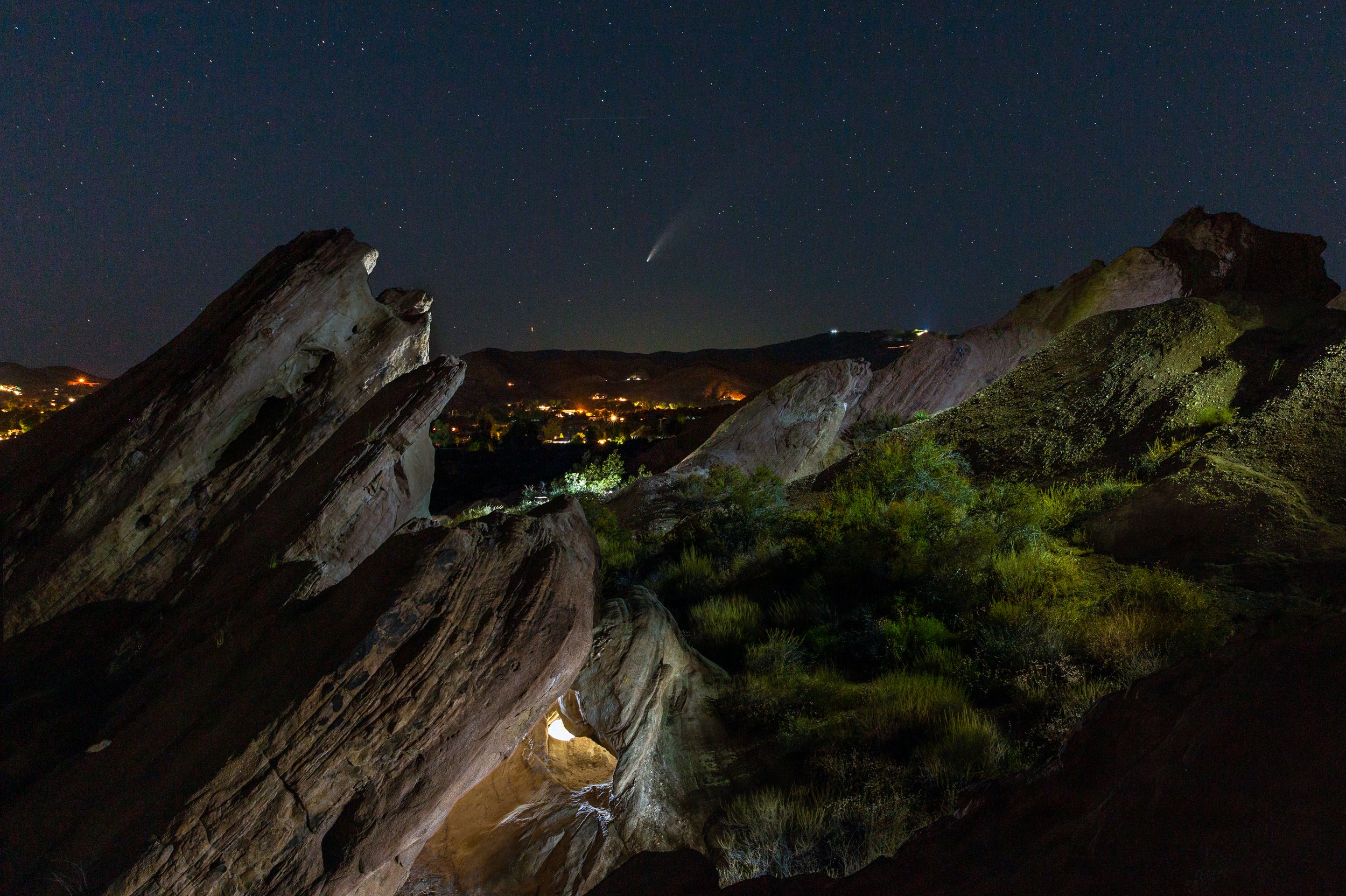 Nighttime landscape featuring large rocky formations with a distant town illuminated below, a starry sky, and a visible comet or bright celestial object.