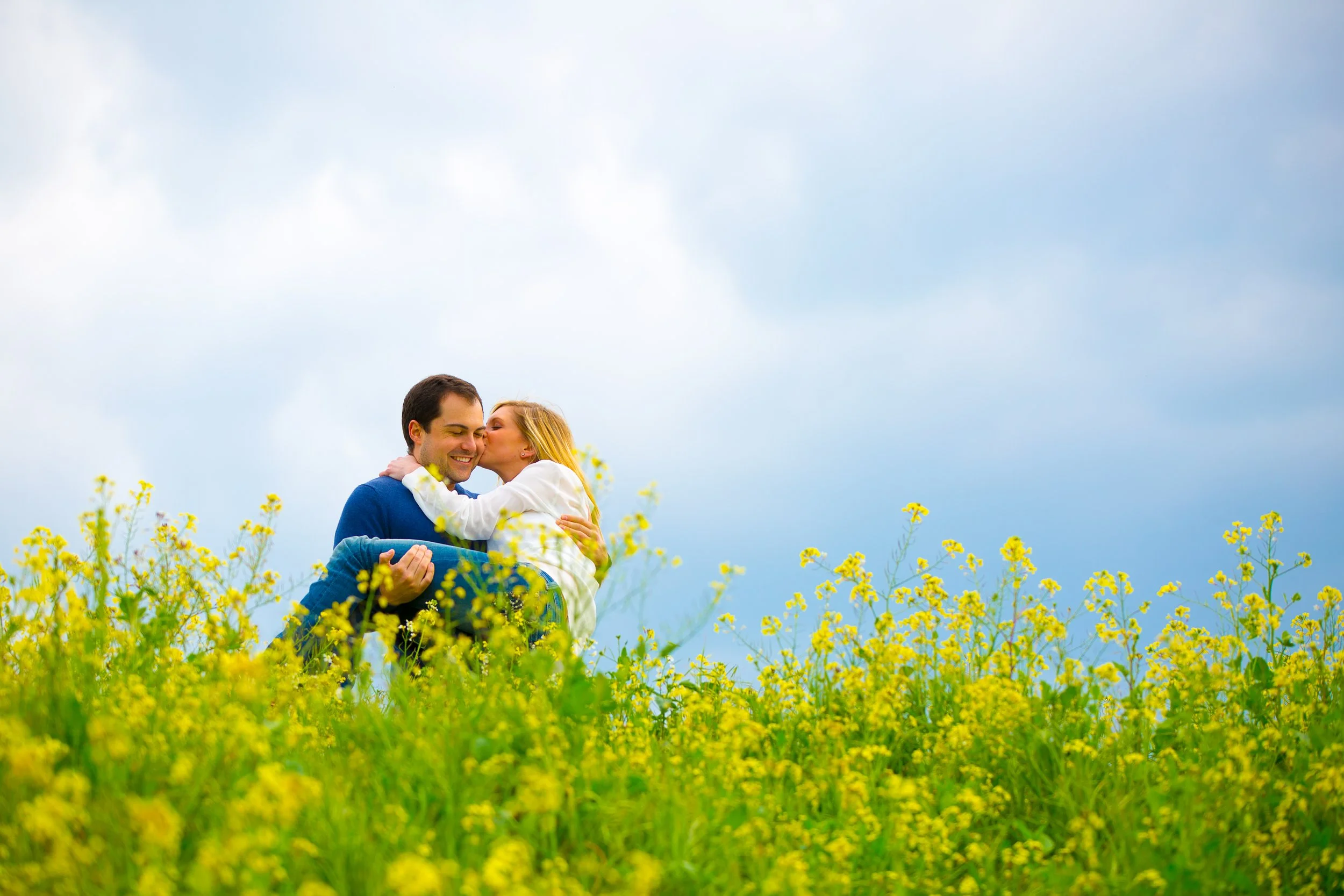A couple in a field of yellow flowers, with the woman kissing the man on the cheek under a cloudy sky.