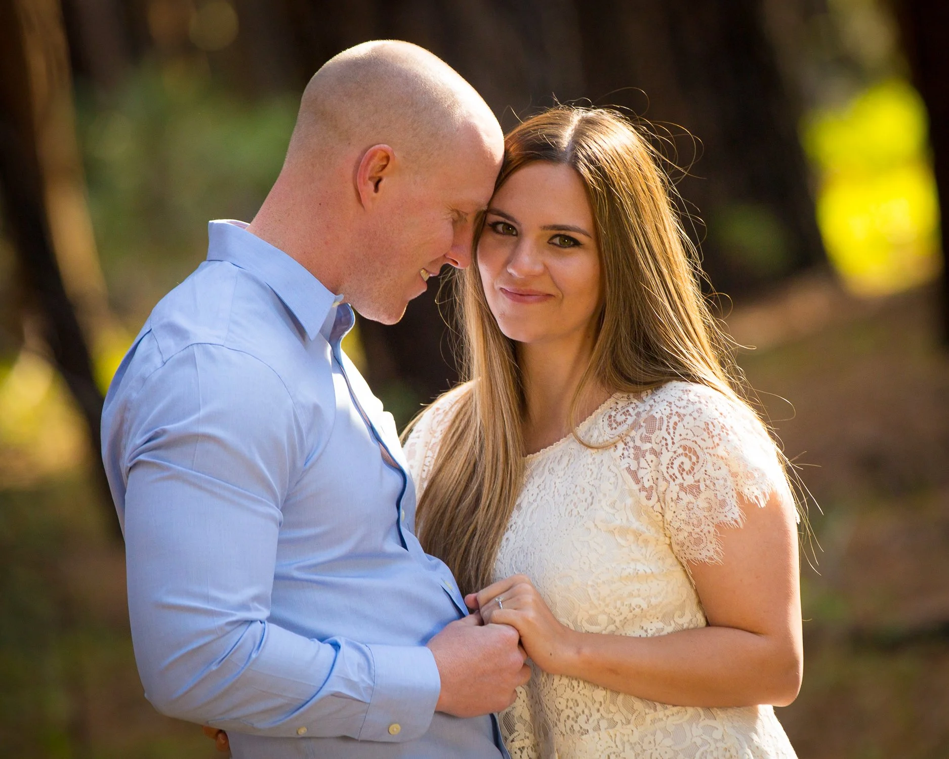 A couple standing outdoors, holding hands and smiling. The man is bald, wearing a light blue shirt, leaning his head toward the woman's forehead. The woman has long light brown hair, wearing a white lace dress, looking at the camera.