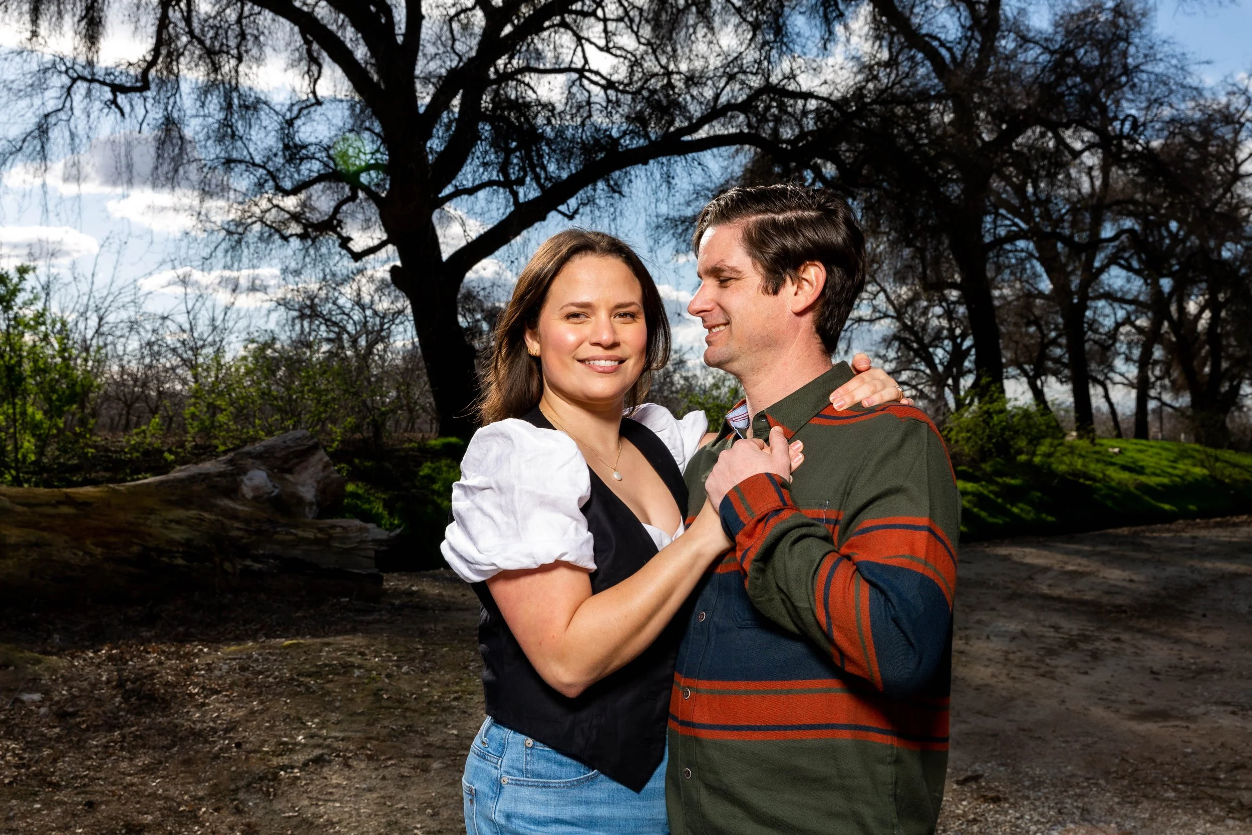 A young couple dancing outdoors near trees with blue sky and clouds in the background.