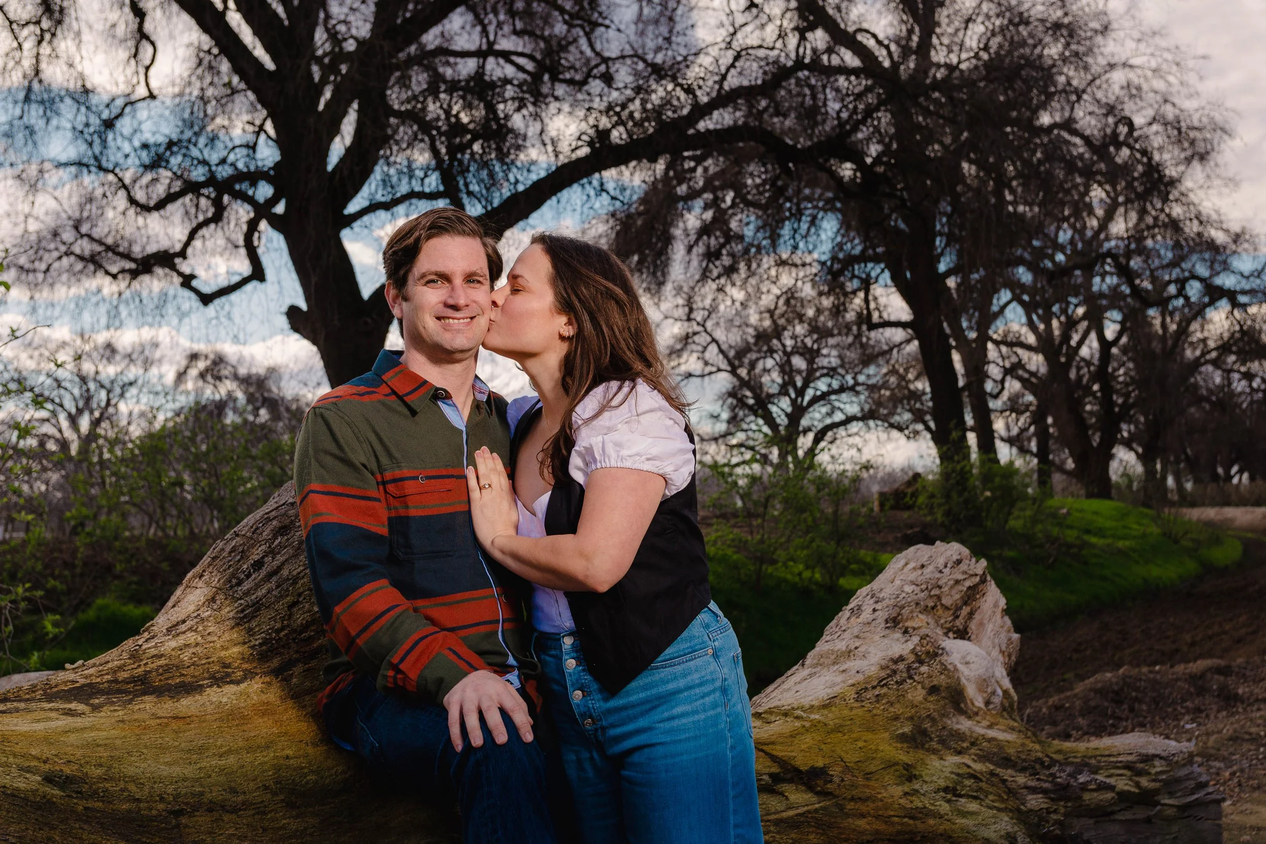 A couple sitting on a fallen log in a park, with trees in the background, one woman kissing the man's cheek. It is daytime with partly cloudy skies.
