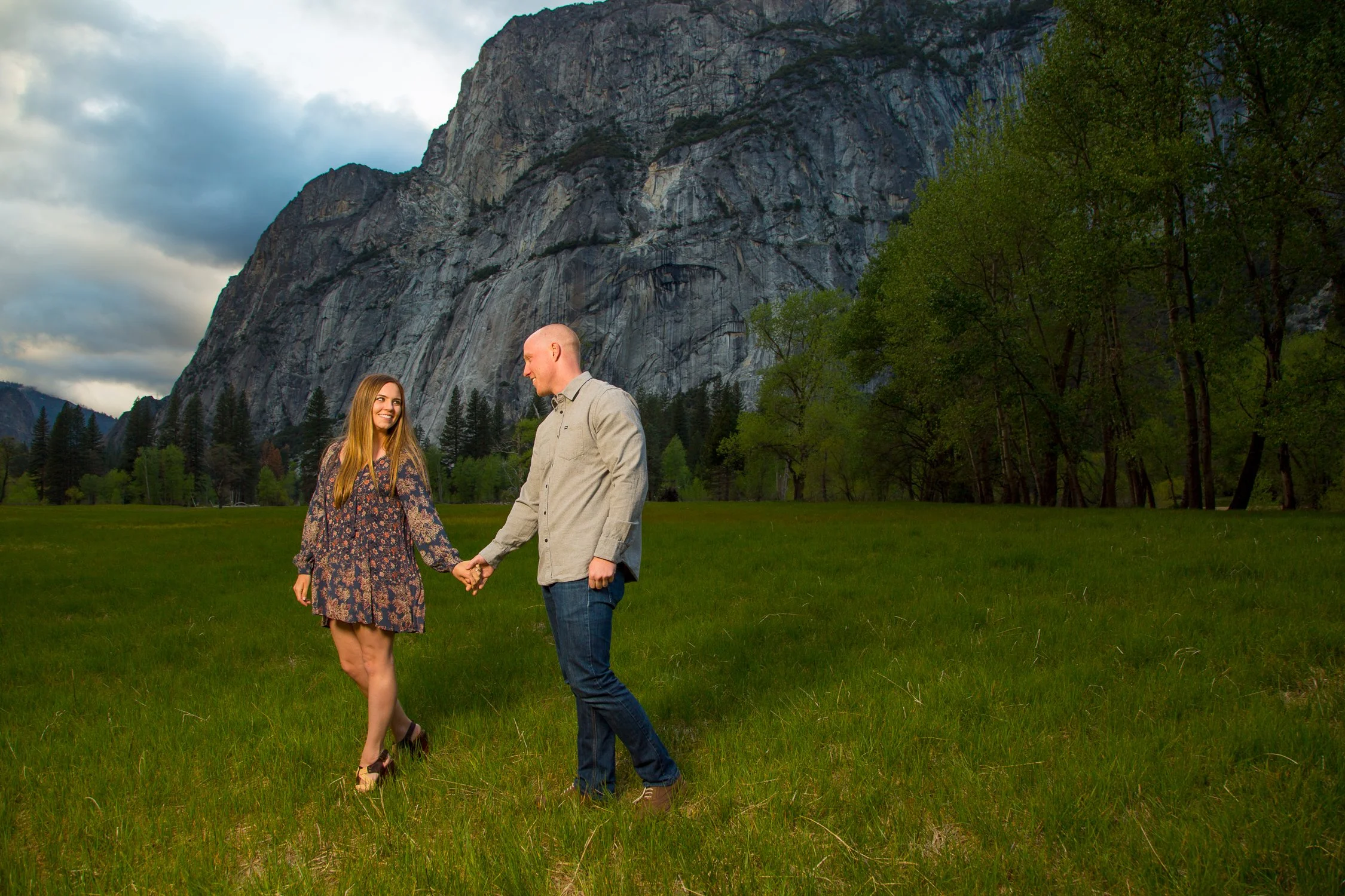 A couple holding hands and smiling at each other in a grassy field with trees and a mountain backdrop.