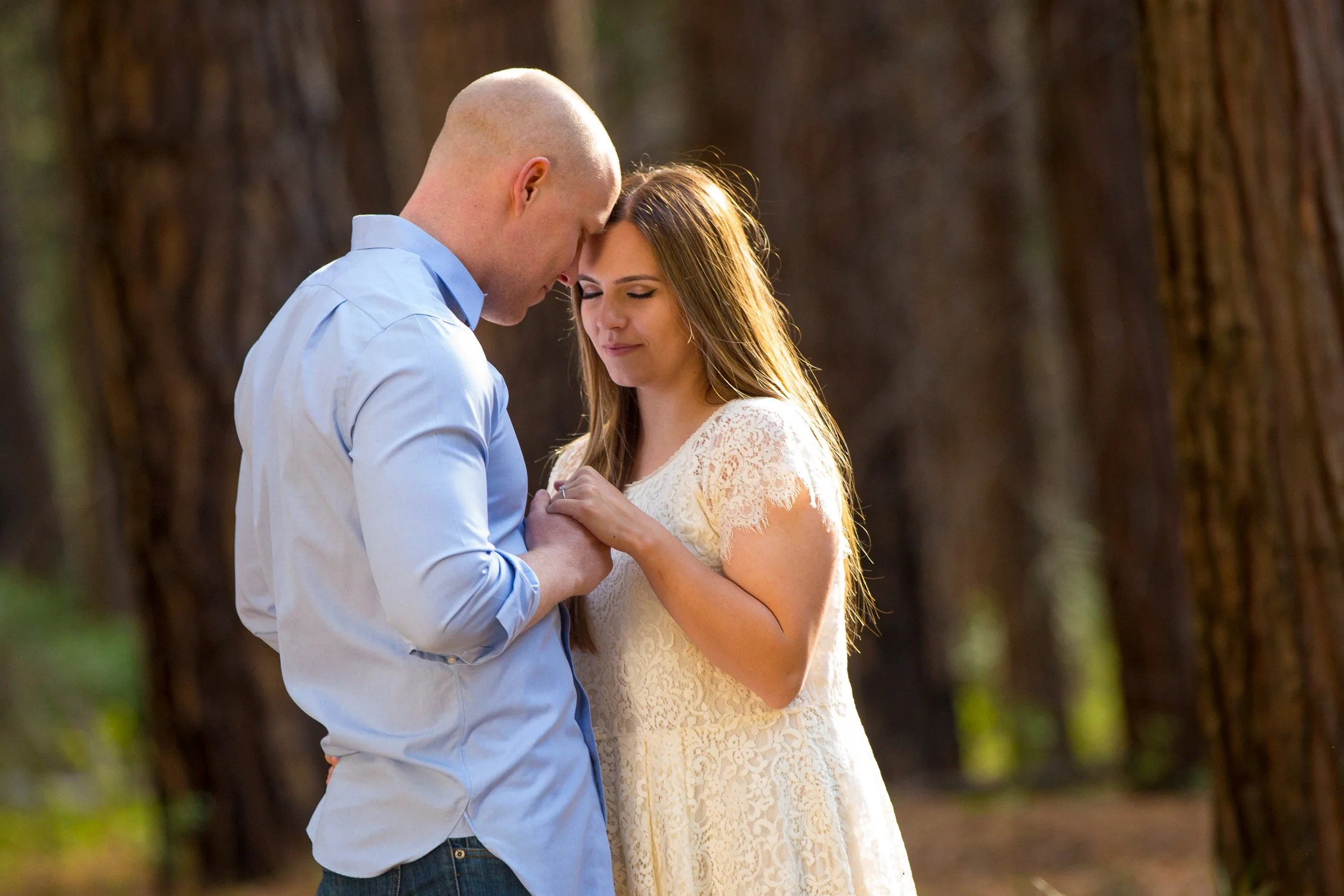 A couple stands close together in a forest, holding hands and touching foreheads with eyes closed, sharing an intimate moment