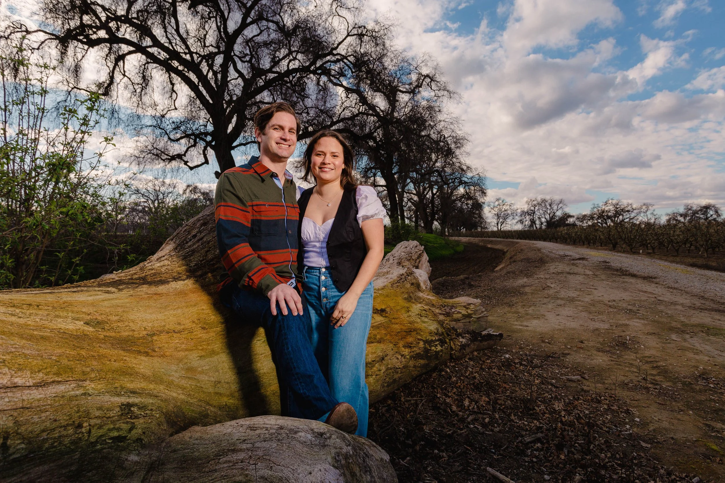 A young couple sitting on a fallen tree in a rural outdoor setting, with a dirt path and leafless trees in the background, under partly cloudy skies.