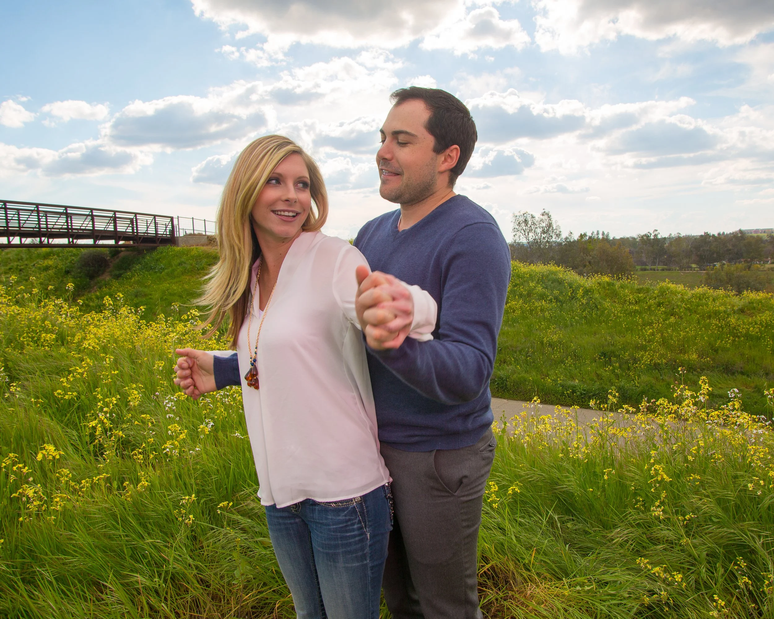 A couple dancing outdoors in a sunny field with yellow flowers, smiling and enjoying each other's company.