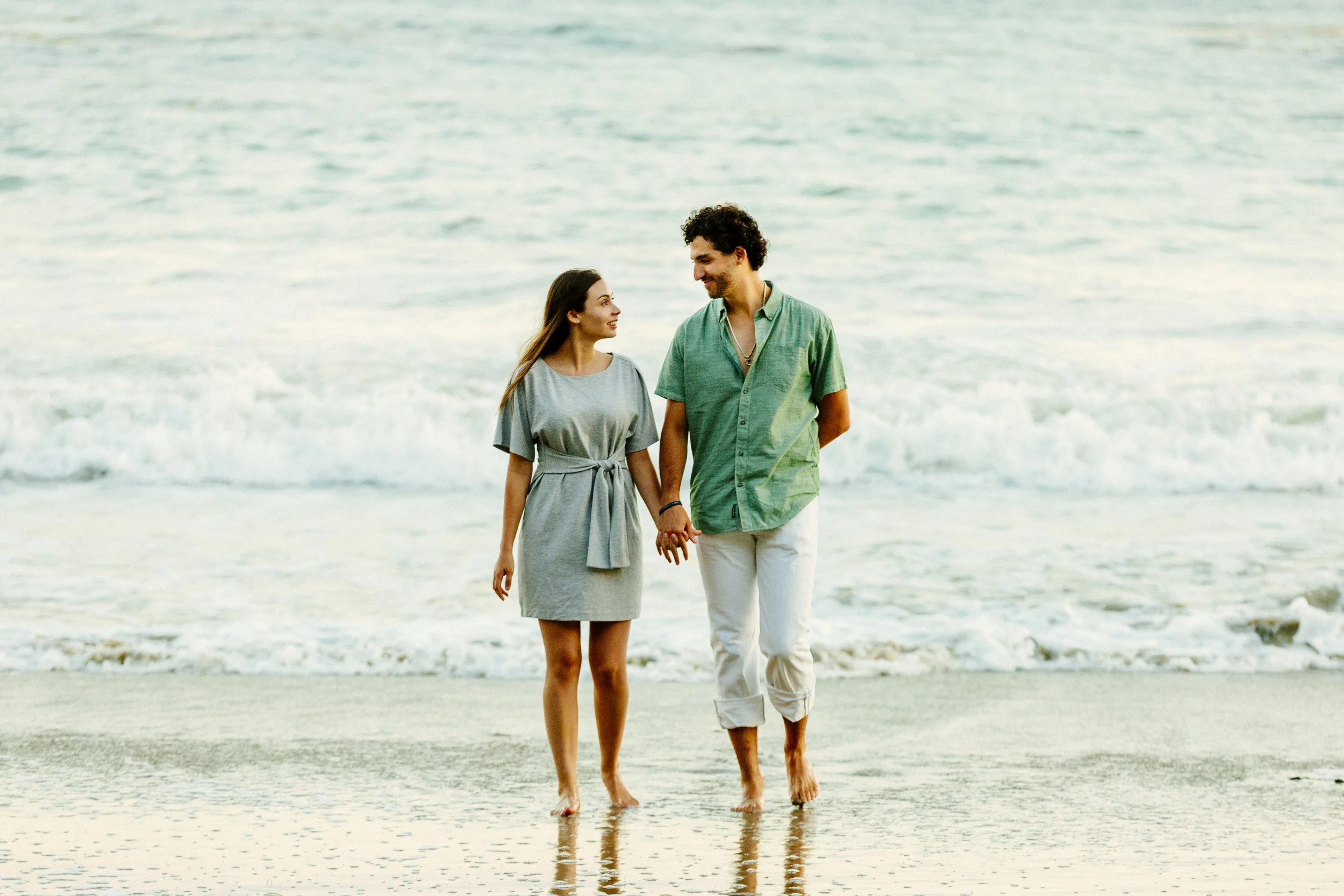 A couple holding hands walking on the beach, smiling at each other.