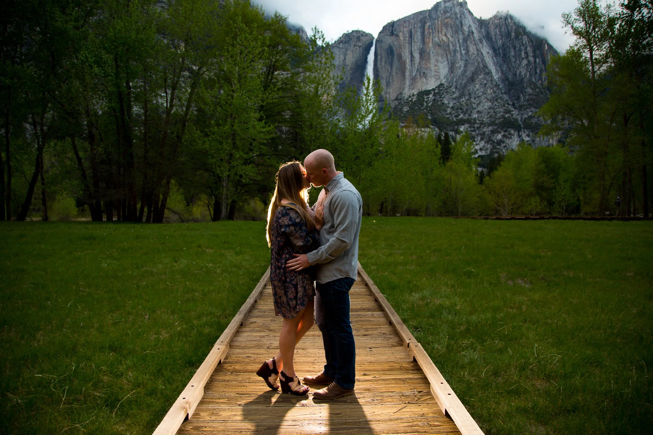 A couple stands on a wooden pathway in a lush green park at sunset, kissing with mountains and a waterfall in the background.