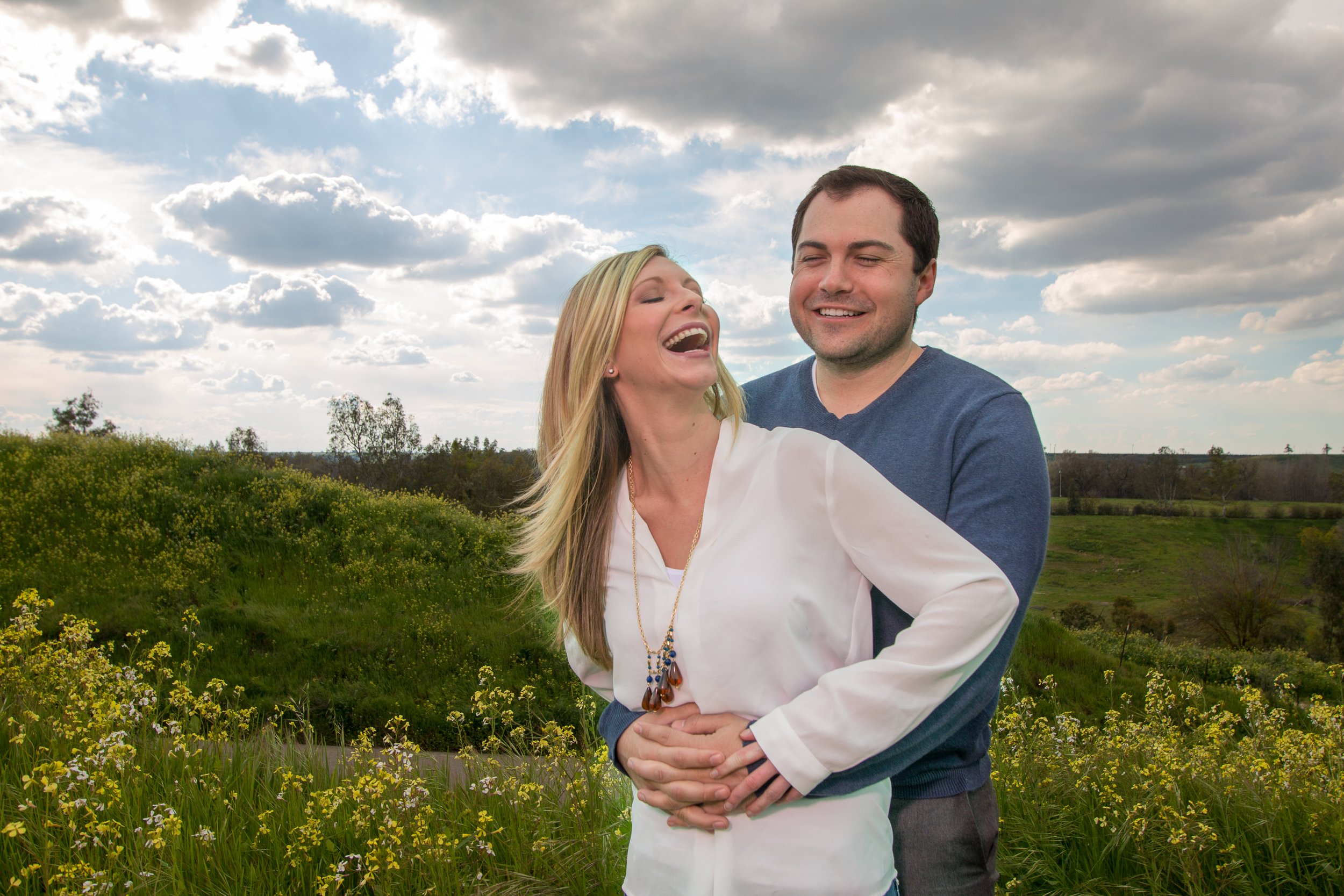 A man and woman embrace outdoors in a field with green grass and yellow flowers, smiling and laughing under partly cloudy skies.