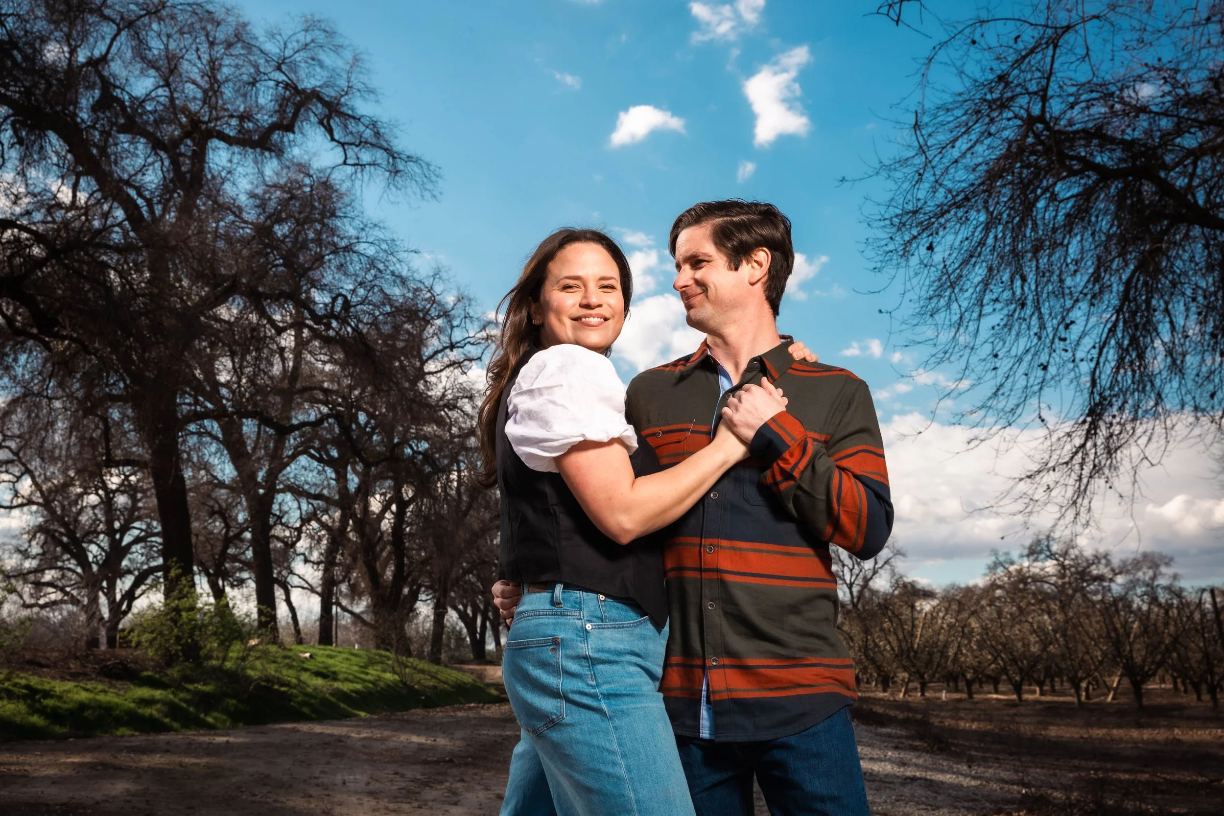 A smiling couple dancing outdoors on a dirt path in a park with bare trees and a blue sky.