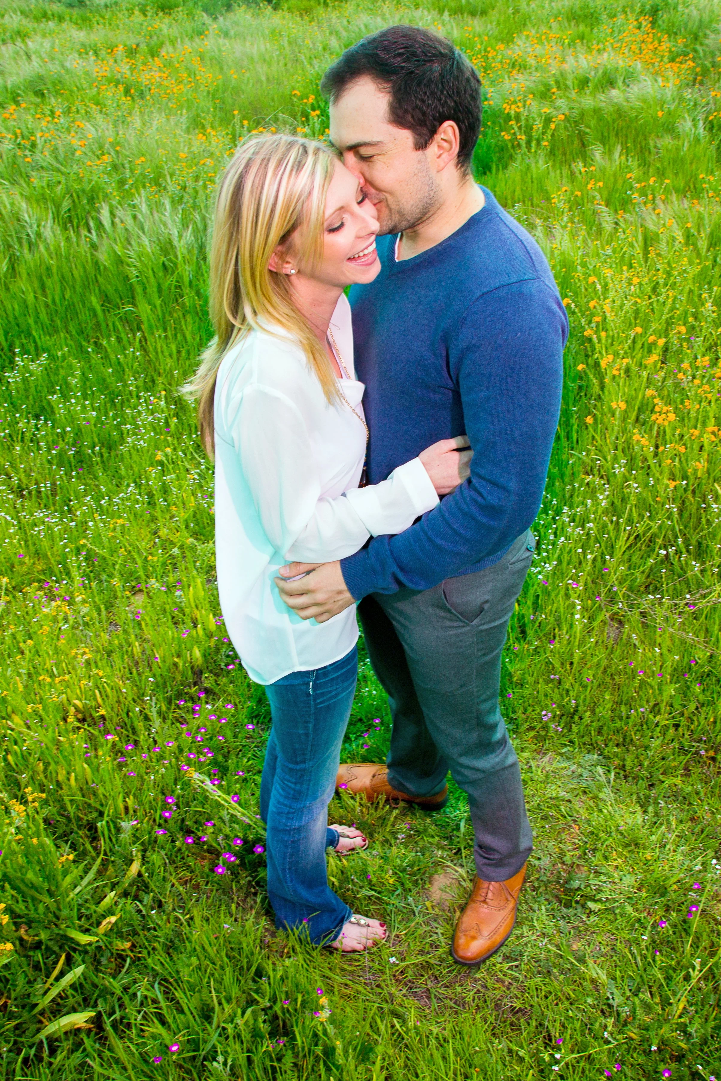 A smiling couple embraces and kisses in a lush green field with yellow and purple wildflowers.