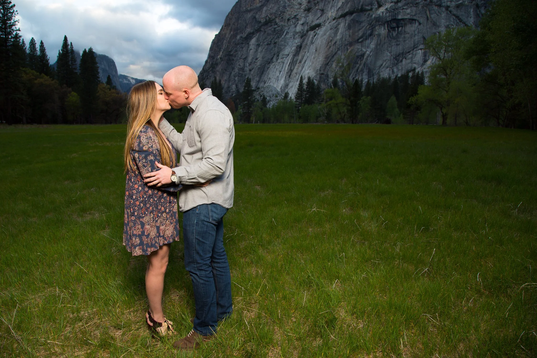 A couple sharing a kiss in a grassy field with tall mountains and trees in the background.