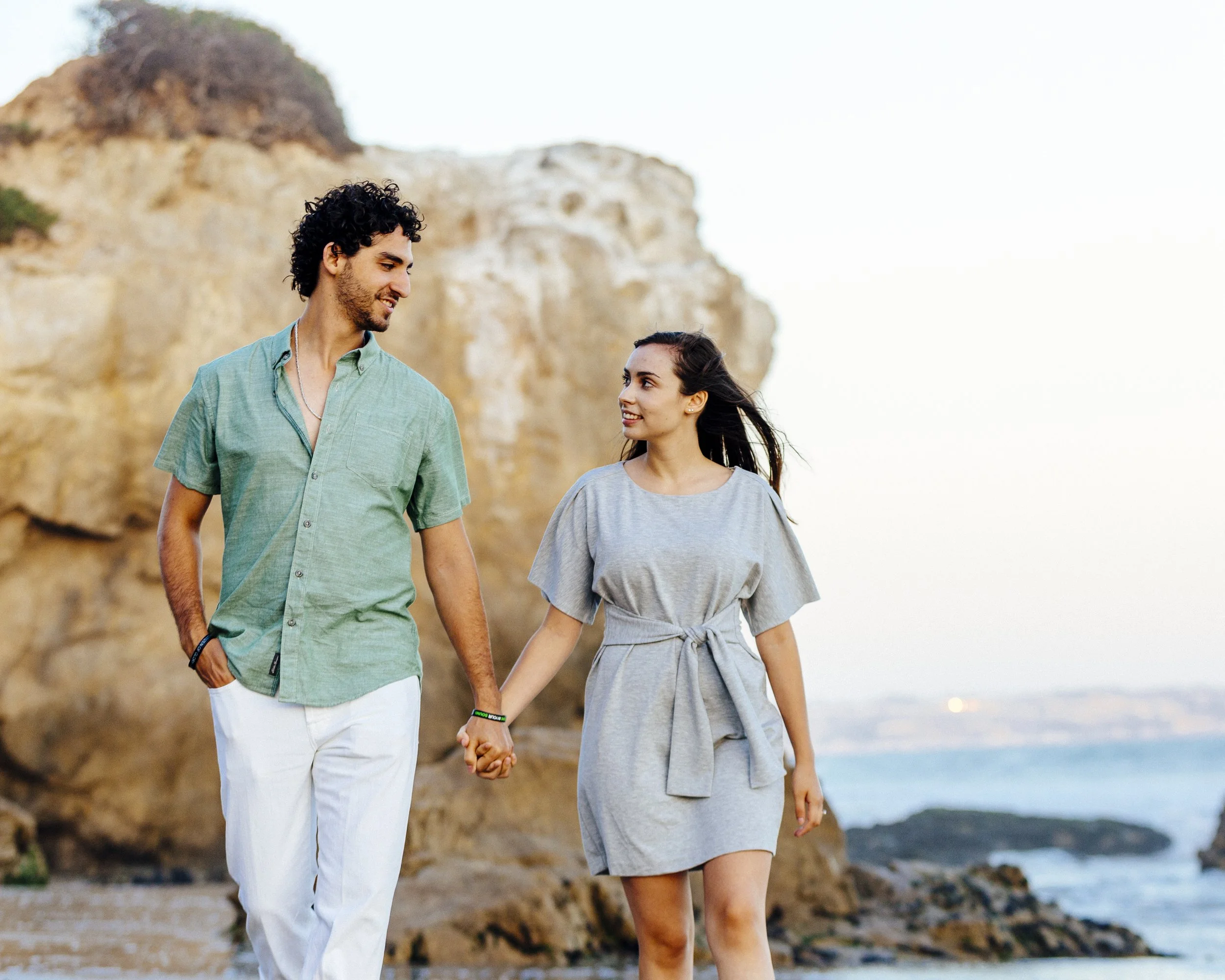 A man and woman are walking hand in hand along a rocky beach, with cliffs and the ocean in the background during sunset.