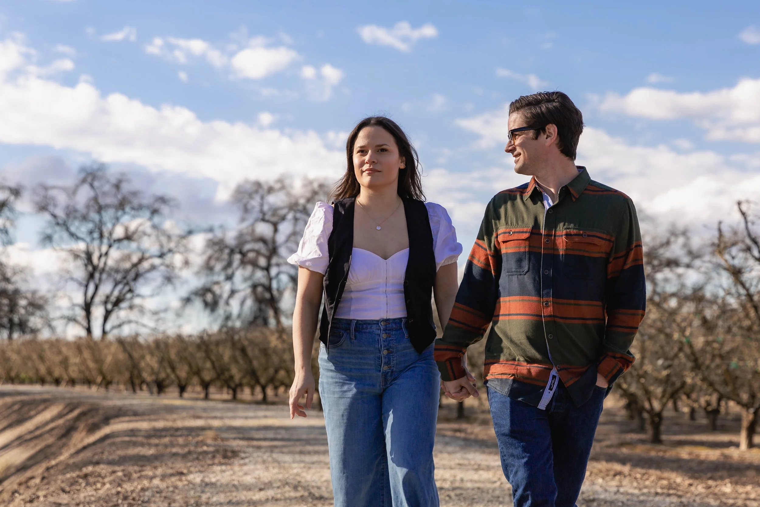 A young woman and a young man walk hand in hand outdoors in a orchard with leafless trees under a partly cloudy sky.