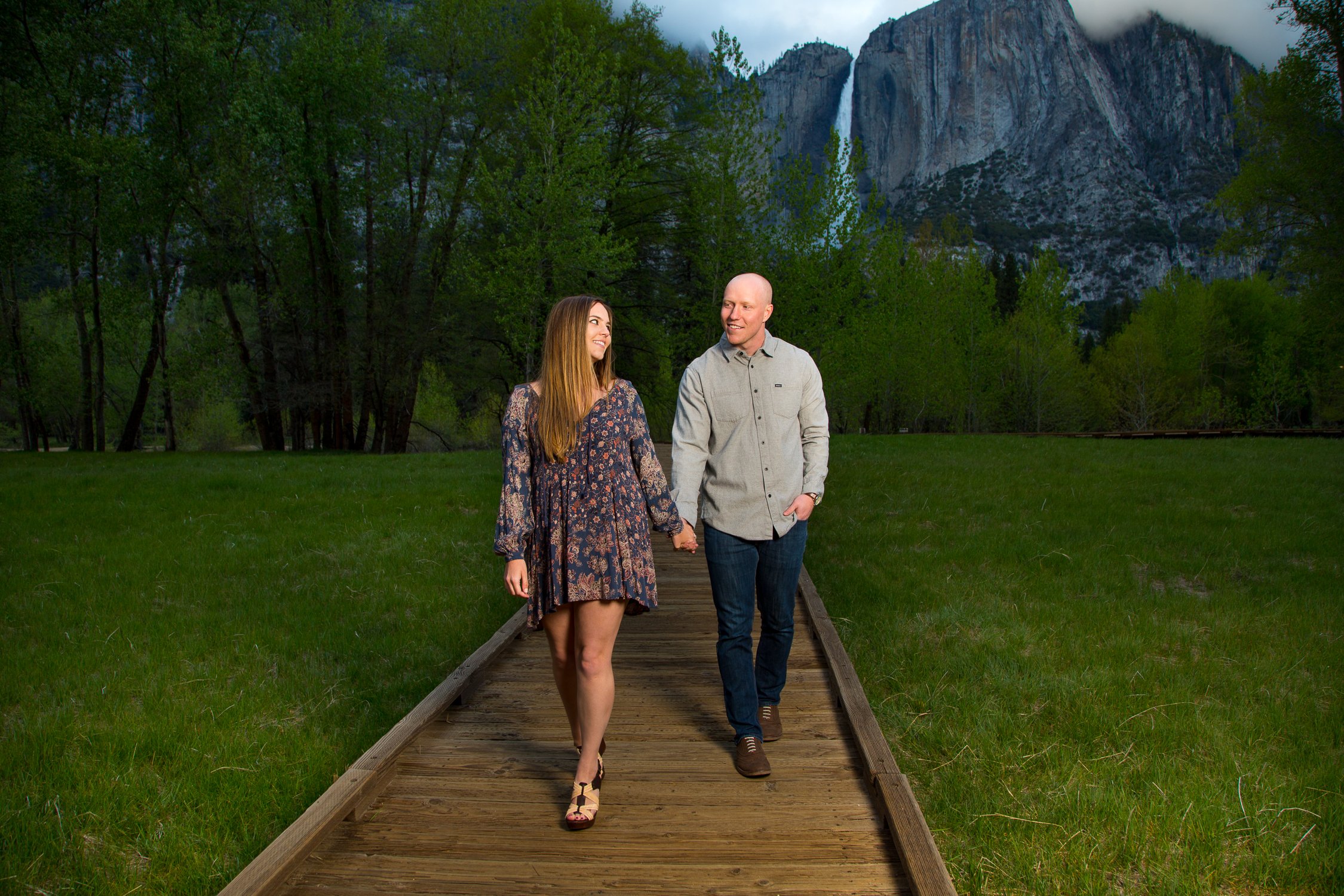 A couple walking hand in hand on a wooden pathway in a park with green grass and trees, with a mountain and waterfall in the background.