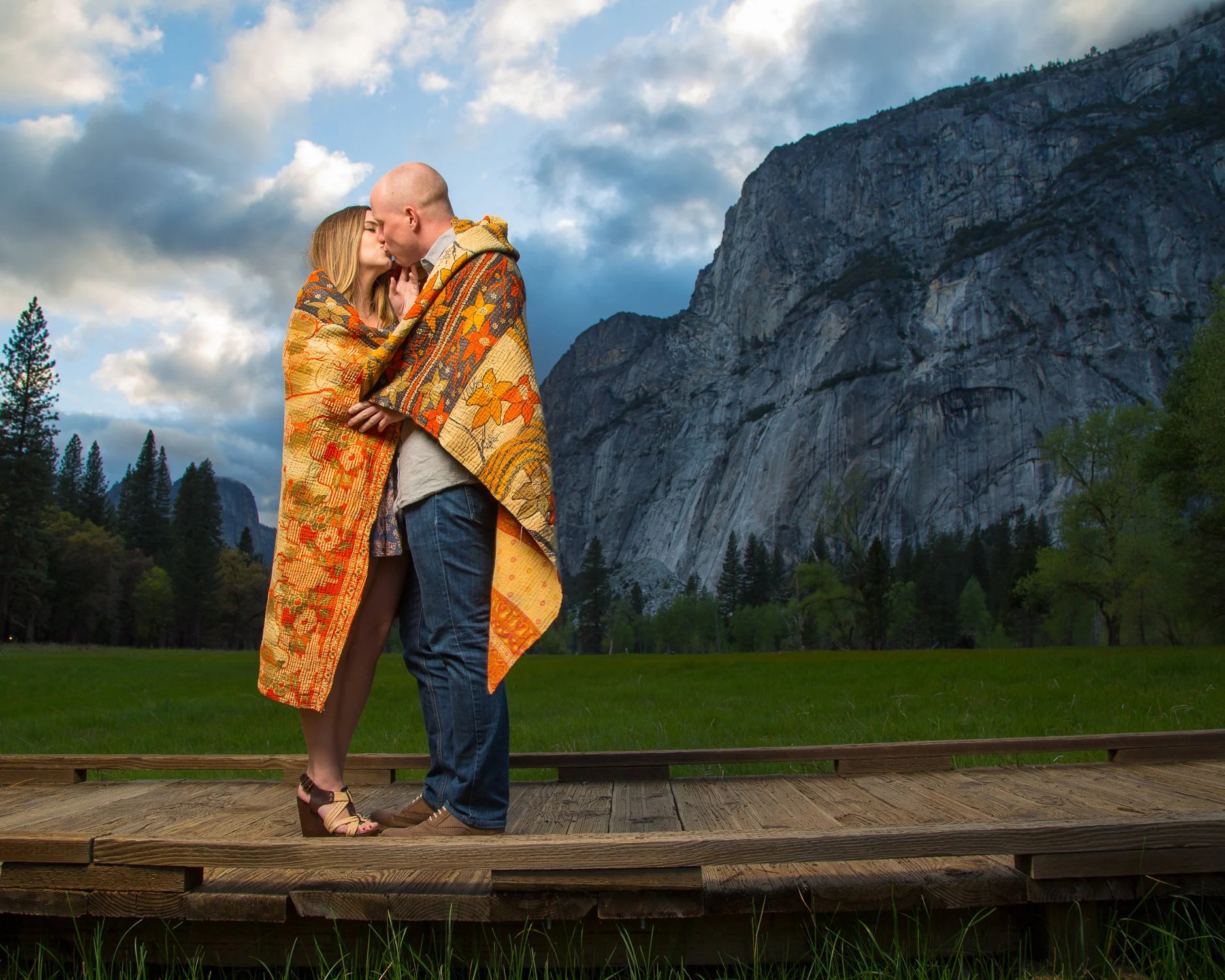 A couple wrapped in a colorful quilt sharing a kiss on a wooden platform outdoors with mountains, trees, and a cloudy sky in the background.